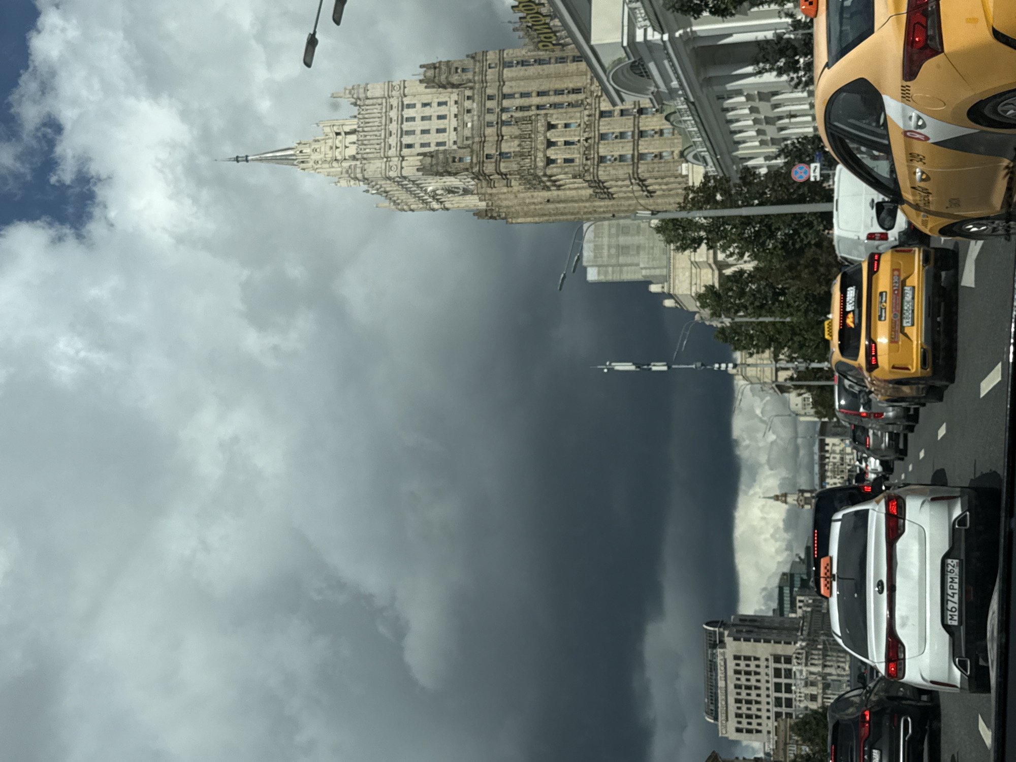 The image captures a street scene in a city under a dramatic sky with dark, ominous clouds. In the foreground, there is heavy traffic with several cars, including white and yellow taxis. One white car prominently displays the license plate "M674PM62". A tall, ornate Stalinist skyscraper dominates the right side of the frame, its facade detailed with many windows and architectural embellishments. The sky is a mix of dark, stormy clouds and patches of lighter, cloudy sky, suggesting impending rain. Other buildings, streetlights, and traffic signals are visible in the background, contributing to a dense urban environment. The overall impression is one of a bustling city street on a day with turbulent weather.