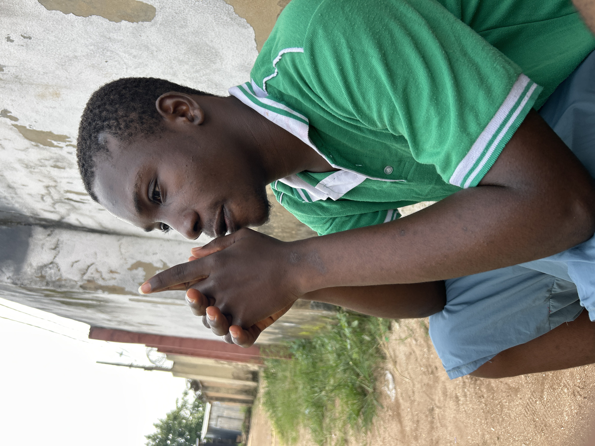 The image shows a close-up, rotated shot of a young Black man sitting outdoors. He is wearing a green polo shirt with white trim on the collar and sleeves, and light blue shorts. His hands are clasped together in front of him, with one index finger pointing. His expression appears thoughtful or concerned. The background consists of a textured wall, some greenery, and what looks like a dirt or sandy ground. In the distance, there are hints of buildings and trees, suggesting an urban or semi-urban environment. The lighting suggests it is daytime, possibly overcast given the diffused light. The scene is located in Ihiagwa, Nigeria. There is no visible text in the image.