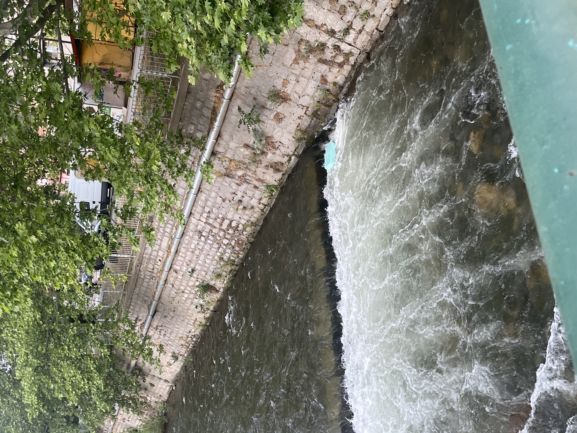 The image shows a river flowing through a stone-lined channel in Blagoevgrad, Bulgaria. The water is turbulent, with rapids on the right side of the frame. Vegetation, including trees and grass, grows along the edges of the stone embankment on the left. A building is visible in the background, partially obscured by trees. A metal pipe runs along the wall of the embankment. The view is from above, possibly from a bridge. A green structure (likely part of the bridge) is visible on the right side.