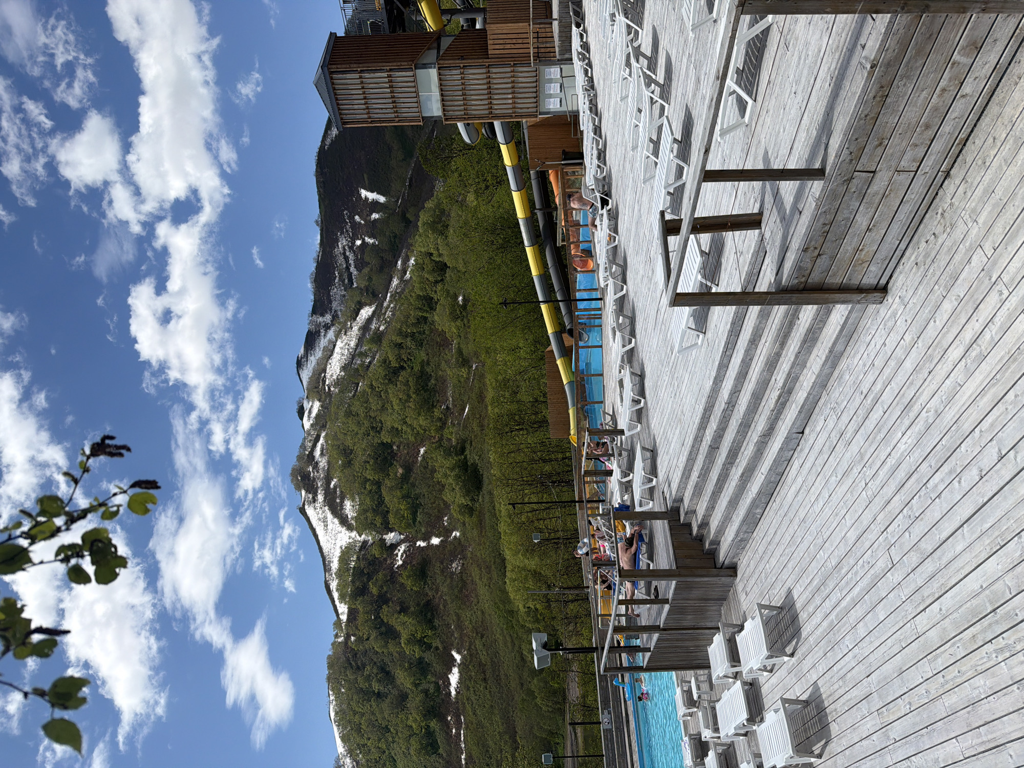 The image depicts a scene at a mountain resort, likely in Paratunka, Russia, given the surrounding landscape. A large wooden deck area is visible, furnished with numerous white lounge chairs, some occupied by people sunbathing or relaxing. A swimming pool with bright blue water is to the left of the deck, with a few individuals swimming or wading. In the background, a towering structure with wooden accents and a yellow and gray water slide is present, suggesting a water park or recreational facility. Beyond the resort structures, a lush green mountainside, partially covered in snow, rises under a bright blue sky with scattered white clouds. The overall impression is of a sunny day at an outdoor recreational area in a mountainous region.