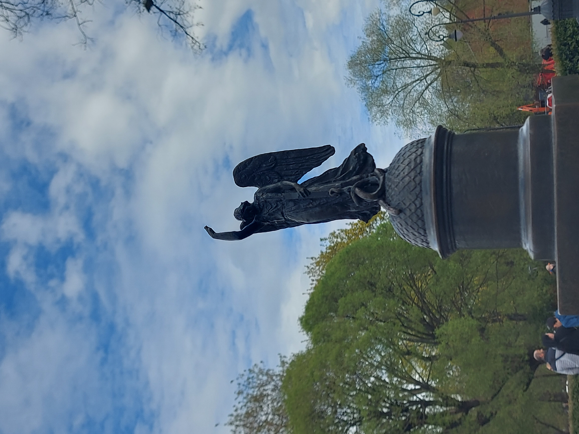 The image depicts the Angel of the Alexander Column in Palace Square, Saint Petersburg, Russia, against a cloudy blue sky. The angel, with wings spread, stands atop the column, its arm outstretched. The column's base, adorned with a decorative acorn, is visible. Trees with green foliage surround the monument. Several people are partially visible at the base of the structure.