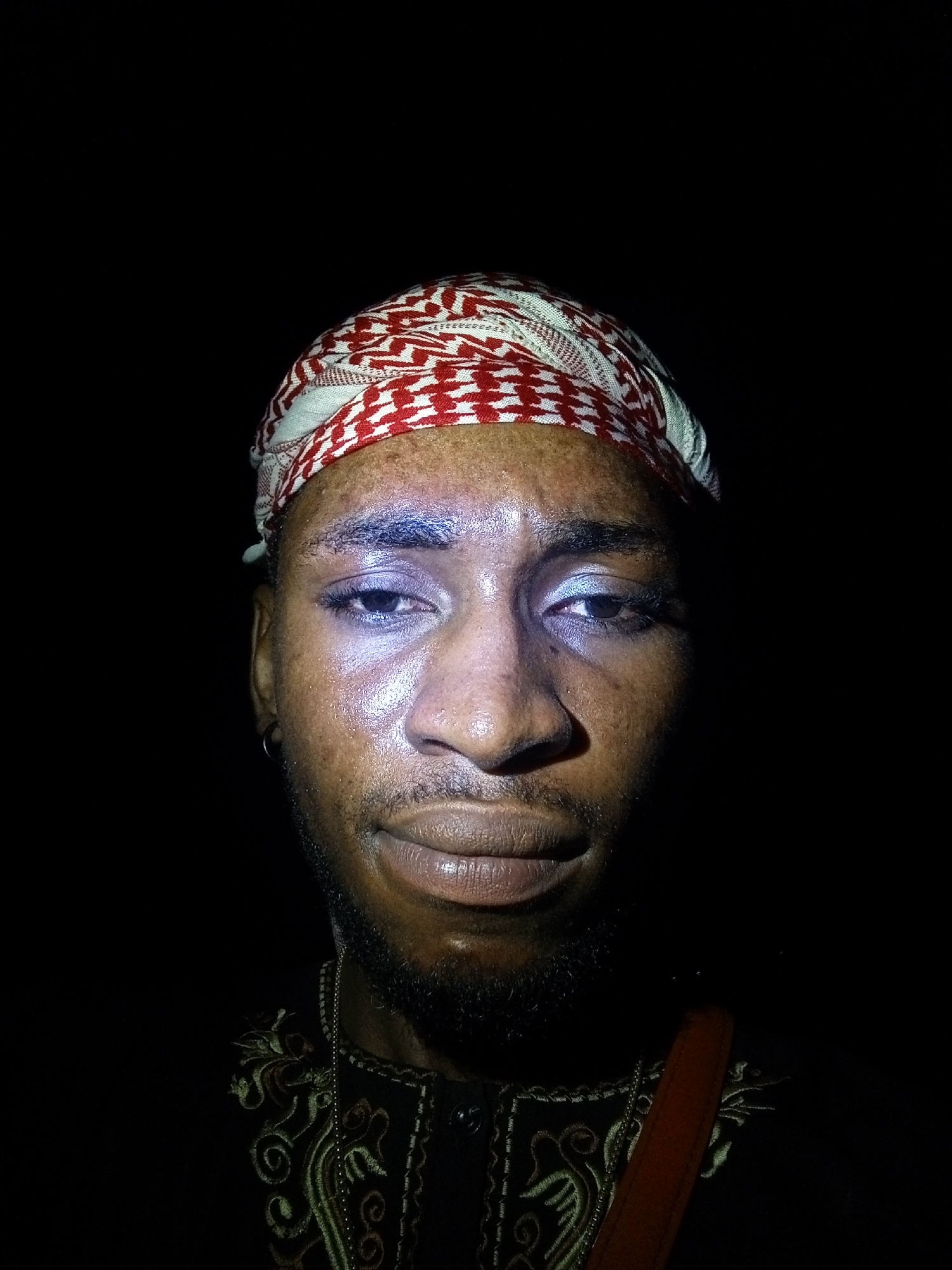 A close-up photograph captures a person against a stark black background. The person is wearing a red and white patterned head covering. Their skin appears to be glistening, potentially due to sweat or oil. A short beard is visible on their face. They are wearing a dark-colored top with intricate gold embroidery and a thin gold chain. A reddish-brown strap is visible diagonally across their torso.
Without additional information, the exact location in Lokoja, Nigeria, cannot be determined. The picture appears to be taken at night or in very low-light conditions.