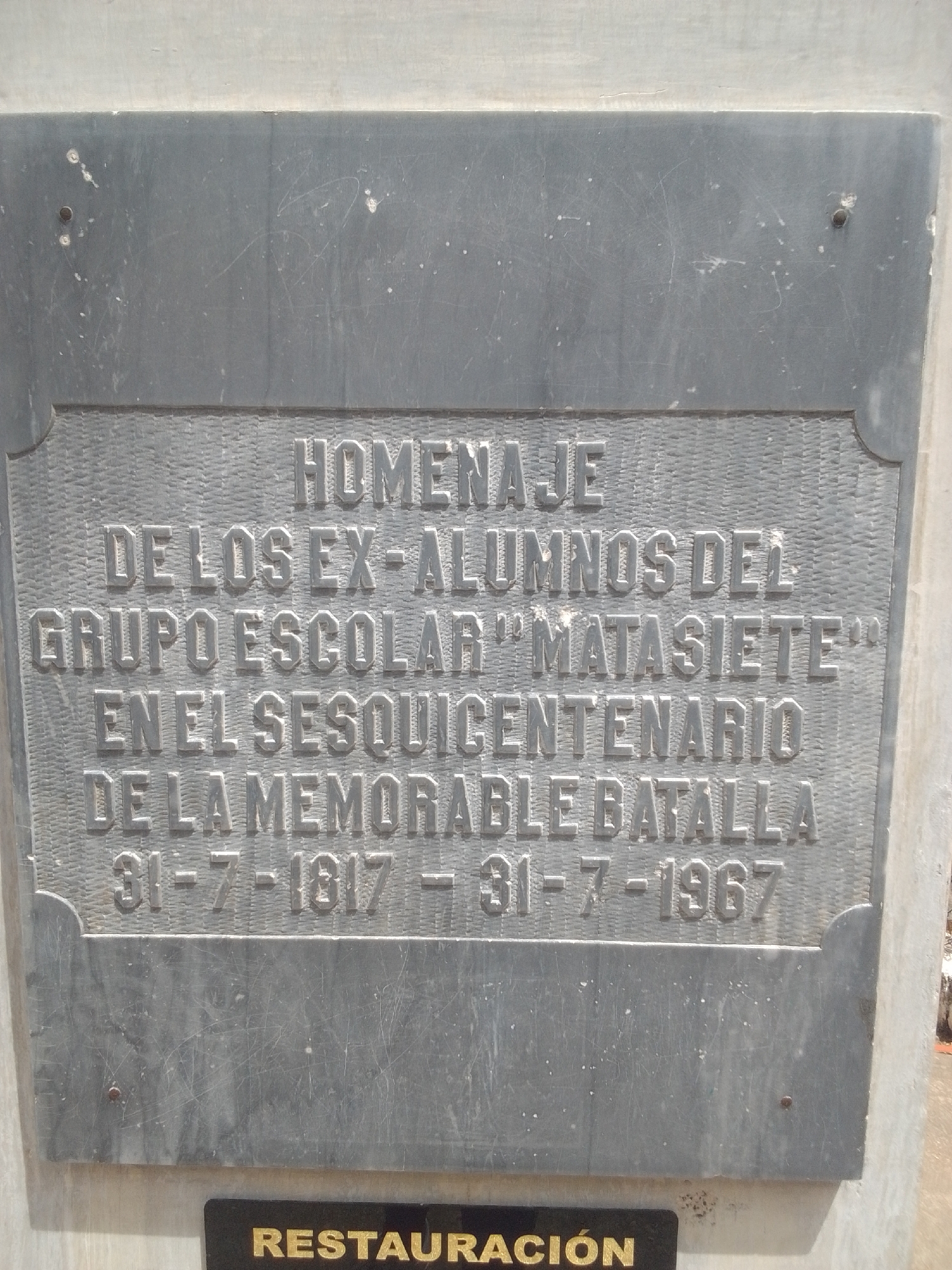This media file features a close-up view of a weathered, dark gray plaque mounted on a light-colored concrete structure. The plaque is engraved with text in raised, silver-colored lettering. The surface of the plaque shows signs of age, with numerous scratches and some white speckling.
The text on the plaque reads: "HOMENAJE DE LOS EX-ALUMNOS DEL GRUPO ESCOLAR 'MATASIETE' EN EL SESQUICENTENARIO DE LA MEMORABLE BATALLA 31-7-1817 – 31-7-1967". Below this main plaque, a smaller black plaque with gold lettering is partially visible, showing the word "RESTAURACIÓN".
The overall scene suggests a historical monument or memorial. The engraving commemorates an event, "La Batalla", which occurred on July 31, 1817, and the plaque was placed 150 years later, on July 31, 1967, by former students of the "Matasiete" school. The context provided indicates this is located in La Tagua, Venezuela. The lighting suggests it is daytime, and the weather appears to be fair. There are no people visible in the frame. The focus is entirely on the inscribed memorial plaque and its details.