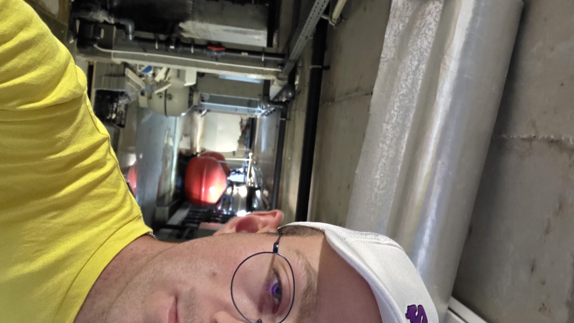A man, partially visible from the chest up and to the right of the frame, wears a yellow t-shirt and a white baseball cap with a purple logo. He is in an indoor industrial or utility space with exposed pipes and concrete ceiling. In the background, there are large red spherical tanks, machinery, and a wet floor. The overall environment suggests a maintenance or technical area. The specific location of Bania, Bulgaria, is not discernible from the image itself.
