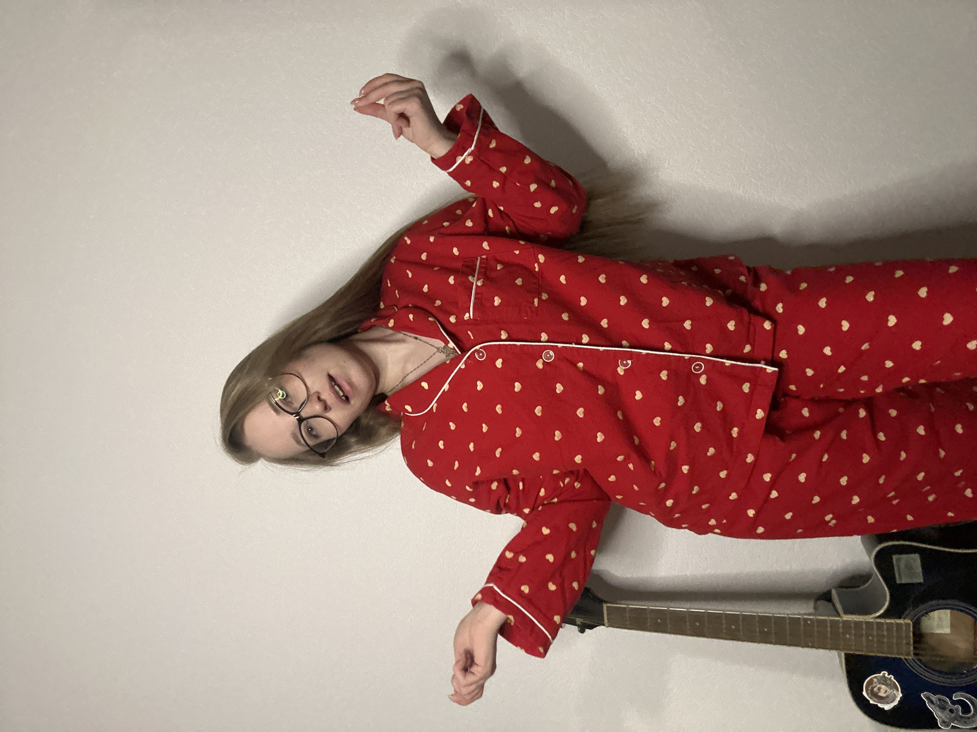 A blonde woman wearing glasses and red pajamas with white hearts lies on a light gray surface next to a black acoustic guitar.