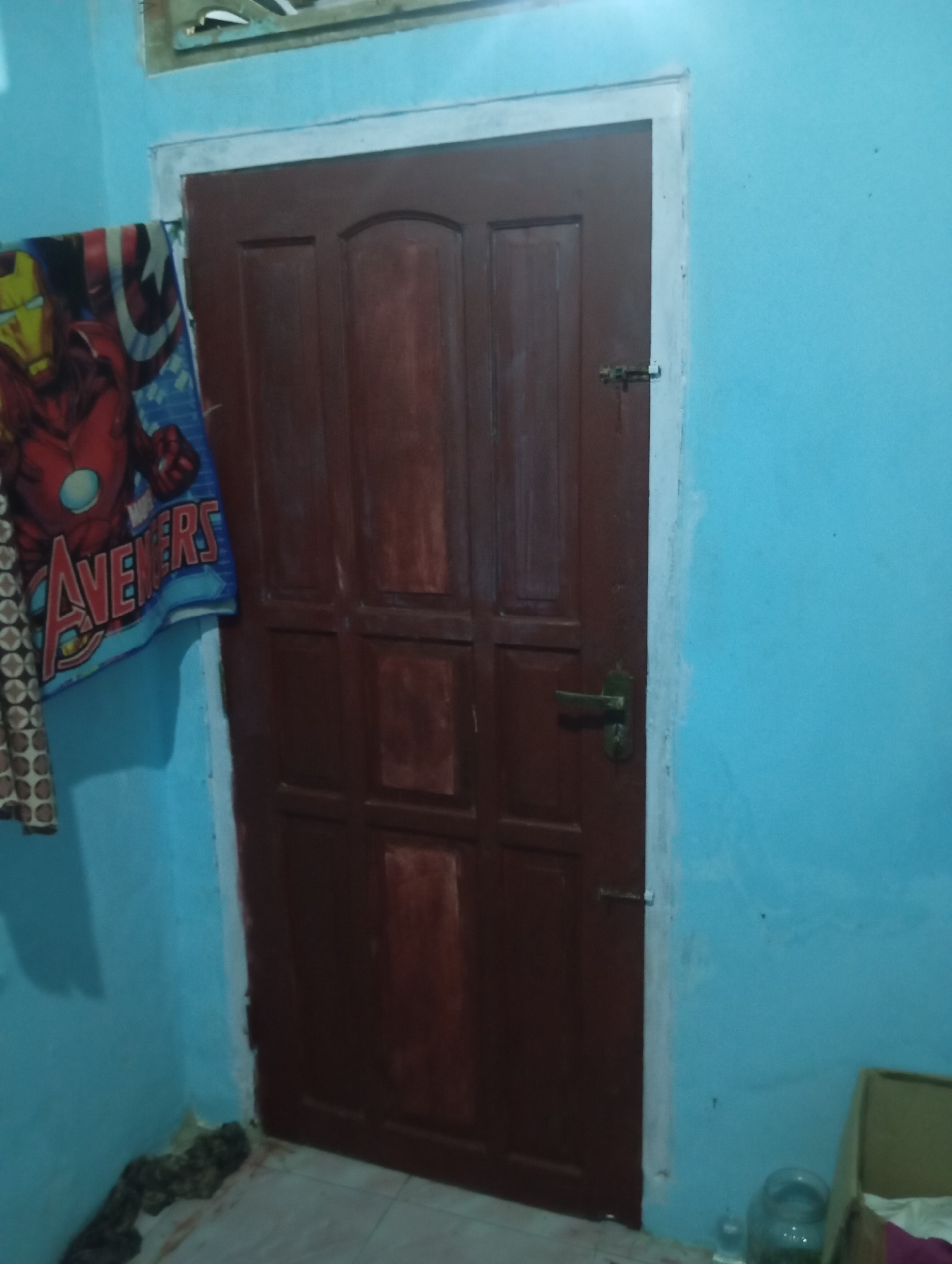 In a room with light blue walls, a dark red wooden door stands closed. The door has several panels and a brass handle and latch. To the left of the door, a blanket featuring Marvel's Avengers characters, including Iron Man and Captain America's shield, is draped over a hook or protrusion. The floor appears to be tiled. In the lower right corner, a glass jar and a cardboard box are visible.