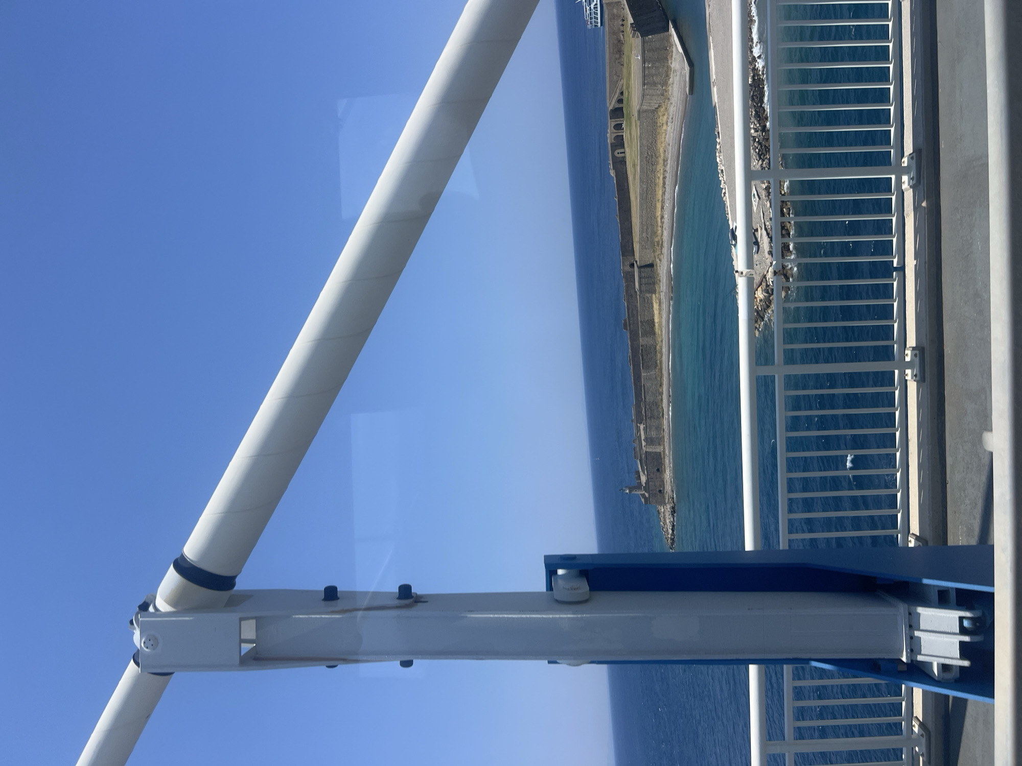 The image captures a view from what appears to be a bridge or ferry, looking out over the sea towards a fortified coastline. In the foreground, large white and blue metal structural elements dominate the left side of the frame, suggesting the vantage point is on a man-made structure. A white railing with vertical bars runs horizontally across the bottom of the image, separating the viewer from the sea.
The sea itself is a deep blue, with visible ripples and a lighter turquoise hue near the shore. On the coastline, a substantial stone fortress with a lighthouse stands prominently. The fortress appears to be old, with a weathered stone construction and arched openings visible. A small beach area is situated in front of the fortress, with some rocky outcrops and what might be a small boat or debris.
Further in the distance, a modern white boat with a blue stripe is visible in the water, suggesting ongoing activity in the area. The sky is a clear, bright blue. The overall scene suggests a sunny day in a coastal location. This view is consistent with the location in the Municipal Unit of Antirrio, Greece, which is known for its bridge and surrounding historical fortifications.