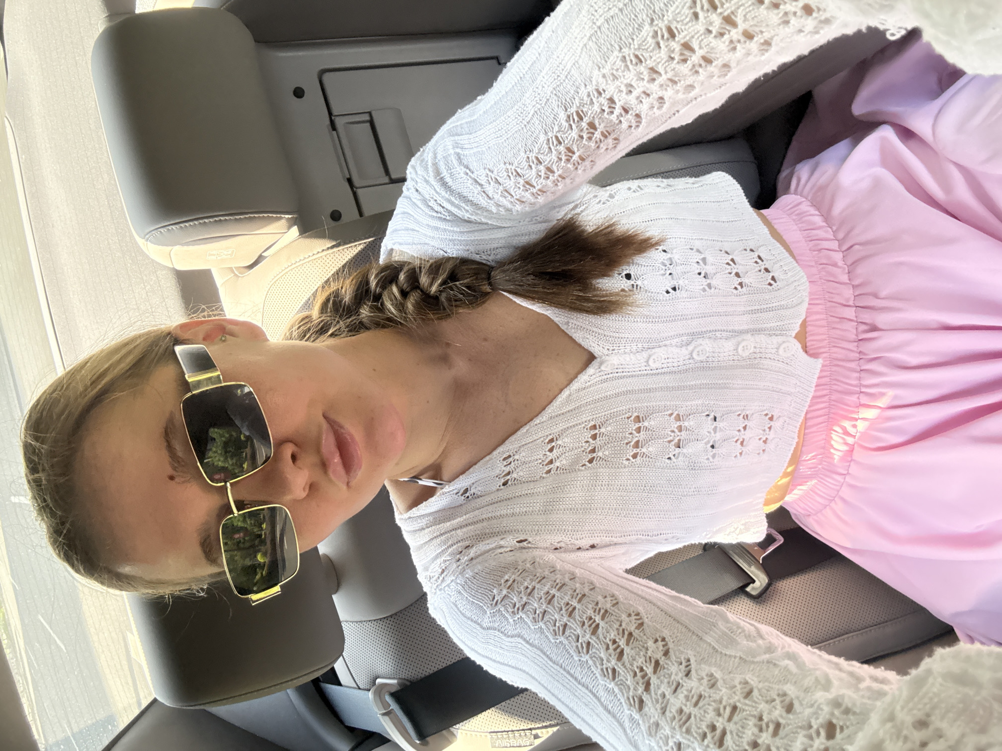 A young woman with blonde hair in a braid is seated in the back of a car, wearing a white crocheted cardigan over a bikini top and a pink skirt. She has on gold-rimmed sunglasses. The car interior, including gray leather seats and a seatbelt, is visible. The scene is outdoors, with reflections of trees and sky in her sunglasses. The image does not contain information about the city of Yiwu, China.