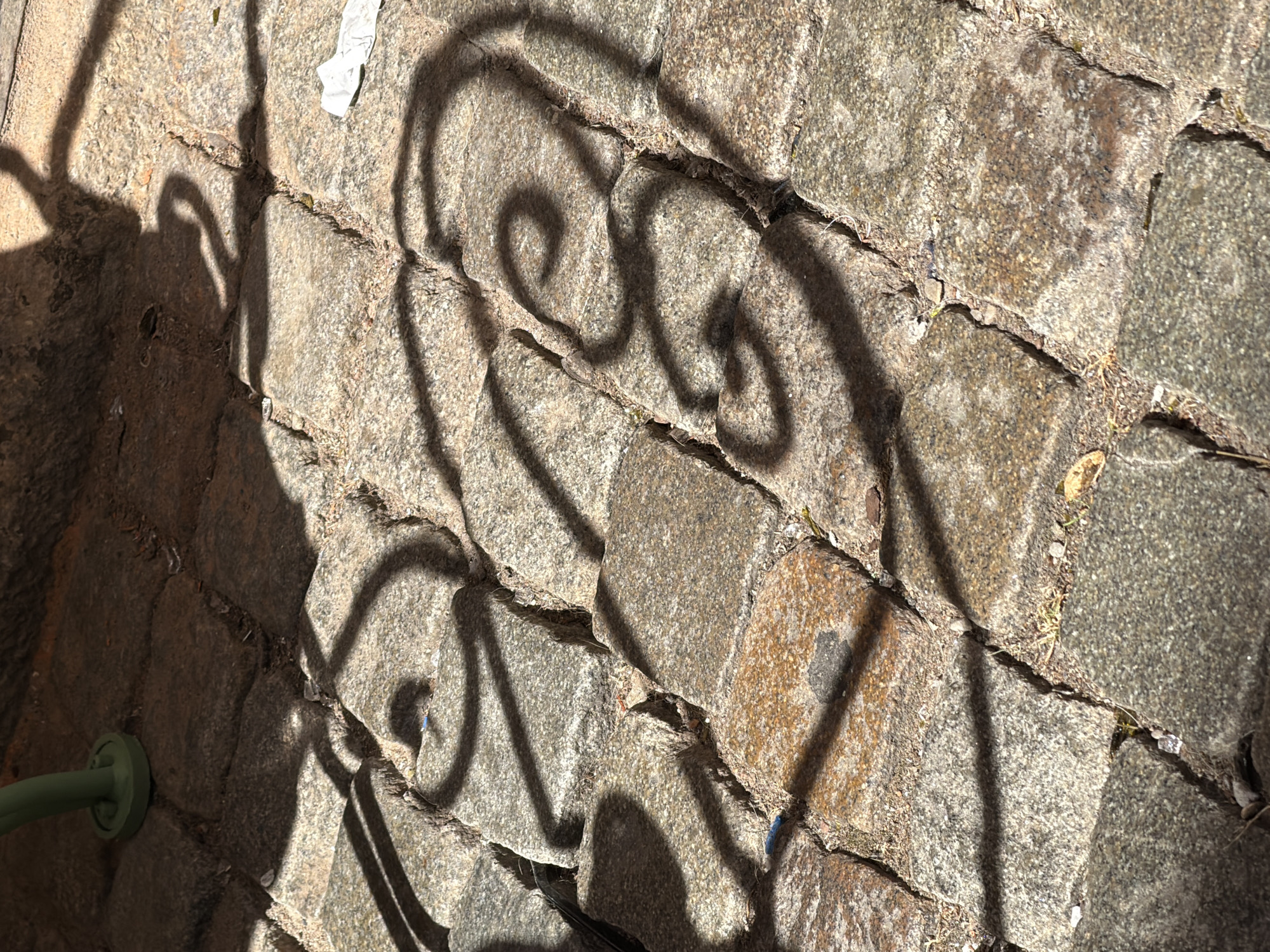 A close-up shot shows the shadow of a decorative metal railing cast upon a cobblestone path. The cobblestones are of varying shades of gray and brown, with visible gaps filled with dirt and small pebbles. The shadow features intricate scrollwork, suggesting a wrought-iron fence or bench. In the upper left corner, a portion of a green metal leg from a piece of furniture is visible, its base resting on the cobblestones. A small piece of white paper lies on the path to the right. The scene appears to be outdoors, likely in a public space within Malmö, Sweden, given the visual cues of cobblestone paving and decorative metalwork often found in European cities.