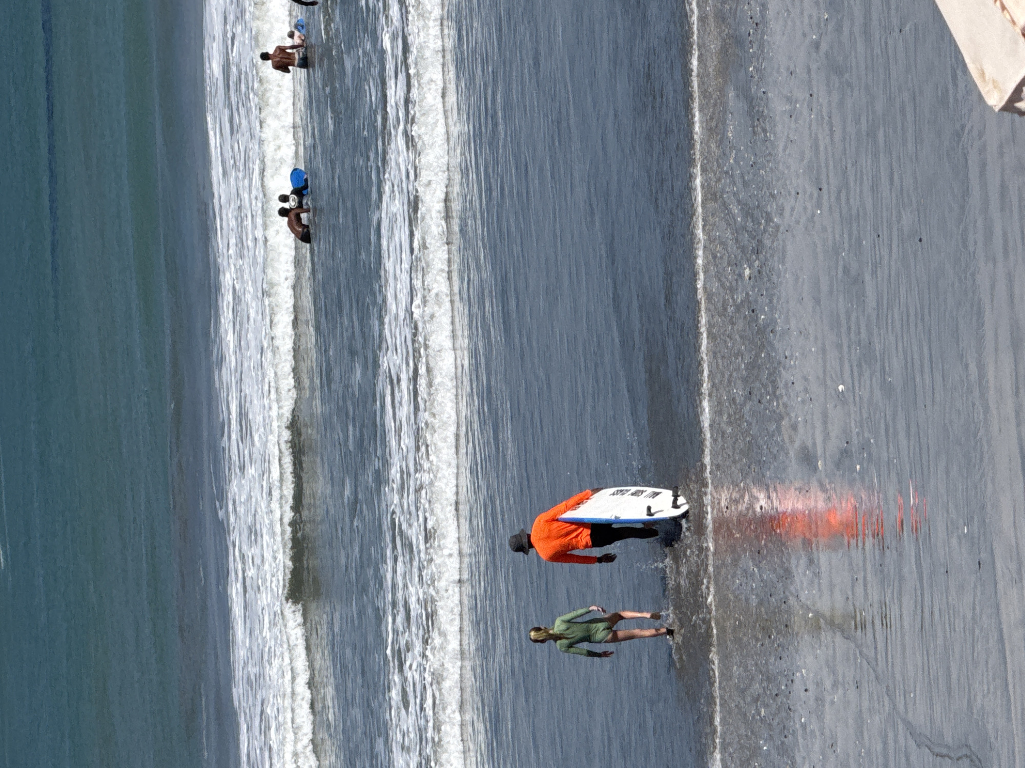 In Kuta, Indonesia, two people are walking into the ocean from the beach. A child in a green wetsuit walks ahead, while an adult in an orange long-sleeved shirt and black shorts follows, carrying a white surfboard. The water is calm with small waves. In the background, several other people are visible in the water, some with surfboards. The sun is shining, creating reflections on the wet sand.