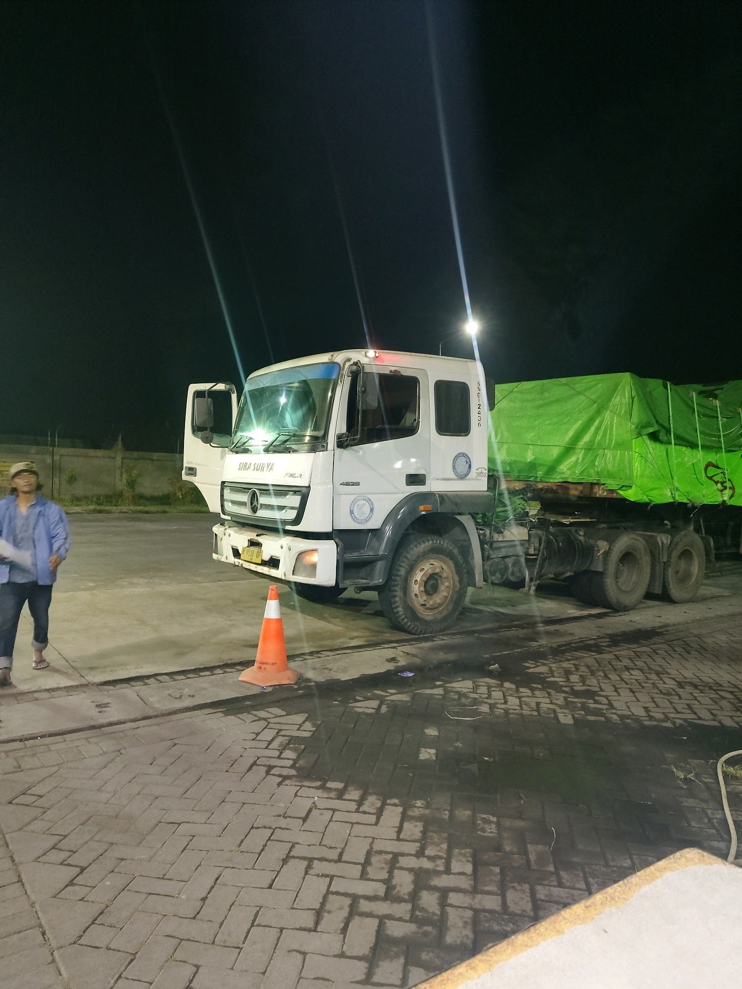 A white Mercedes-Benz Actros truck, license plate B 9538 OF, is parked at night in what appears to be an industrial area in Kedung Sukodani, Indonesia. The truck's driver-side door is open, and its headlights and emergency lights are illuminated. The trailer of the truck is covered with a large green tarp. A person wearing a light blue jacket and dark pants is standing to the left of the truck, facing away from the camera. An orange traffic cone is placed in front of the truck's wheels. The ground is paved with interlocking bricks, and there are puddles of water on the ground, suggesting it may have recently rained. The scene is illuminated by overhead lights, creating distinct shadows.