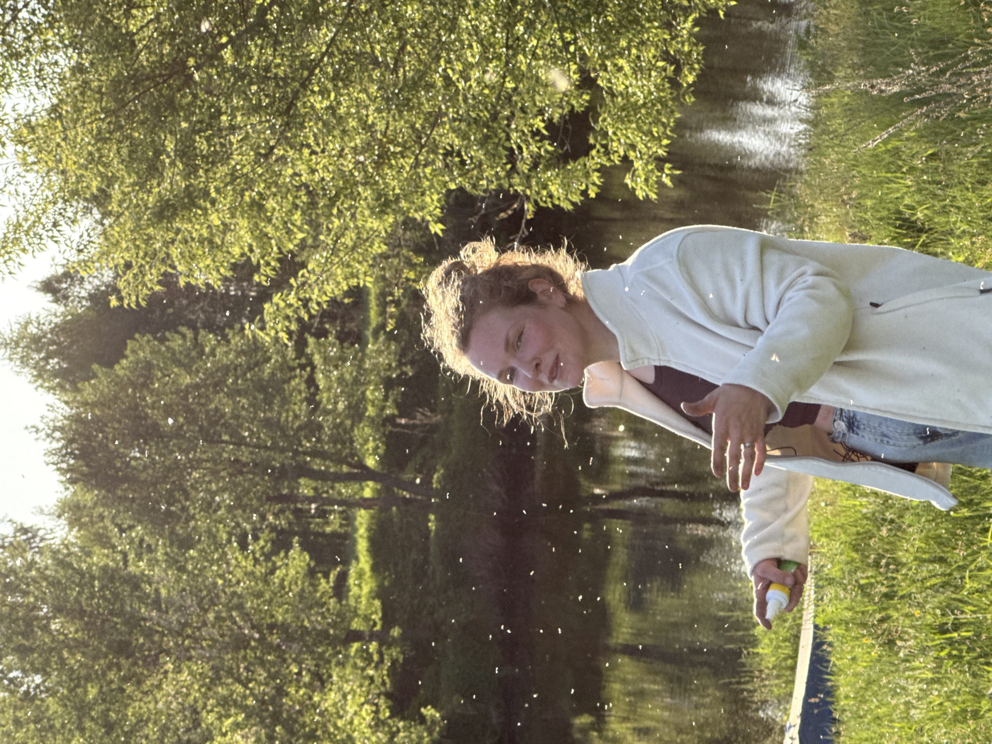 A woman with curly hair stands outdoors beside a body of water surrounded by green trees and grass. She is wearing a white jacket, a maroon top, and blue jeans. In her hand, she holds a yellow and white spray bottle and is apparently spraying something into the air. The location appears to be natural, resembling a park or riverside area. It is impossible to confirm that the location is in Egorii, Russia without additional location data.