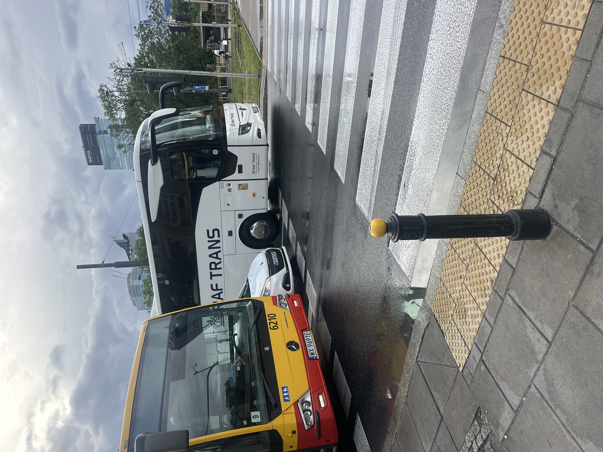 The image shows a street scene in Warsaw, Poland, with two buses stopped at a pedestrian crossing. A yellow and red city bus, with the number 9 visible, is in the foreground on the left. To its right, a white long-distance bus with "RAF TRANS" written on its side is stopped. A white car is positioned between the two buses. The pedestrian crossing is wet, indicating recent rain. The sidewalk has tactile paving for visually impaired pedestrians. In the background, modern buildings are visible, including a skyscraper with the "warta" logo. The sky is cloudy.