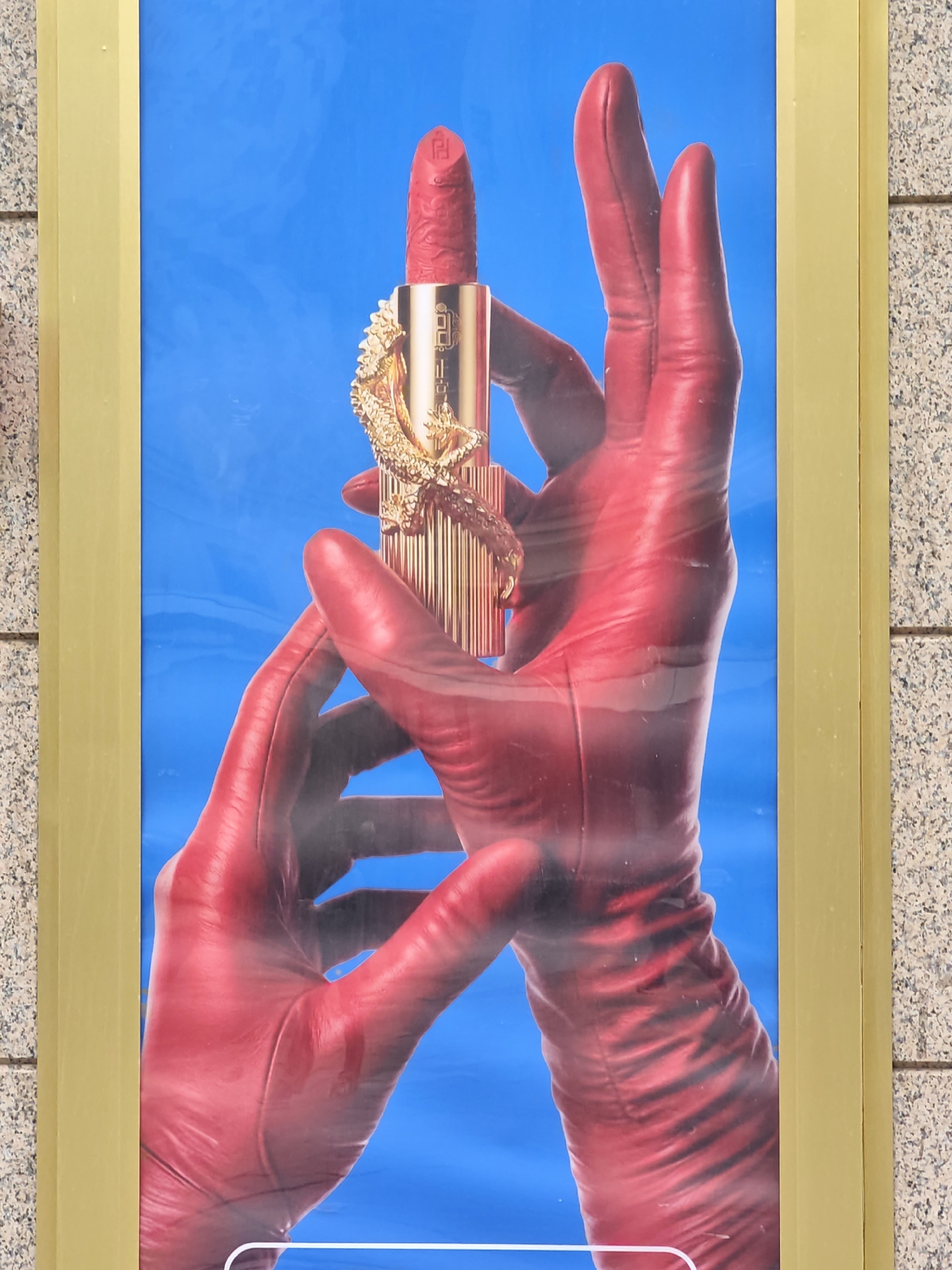 The image is an advertisement poster for lipstick. The poster features a close-up of two red gloved hands holding a red lipstick. The lipstick has a golden dragon design on it. The background of the poster is a vibrant blue. The poster is framed by a yellow border. The poster appears to be on display in a public space, as evidenced by the speckled grey tiled wall in the background which could be a subway station or an exterior wall. The lighting suggests it is daytime. The poster is located in Moscow, Russia.