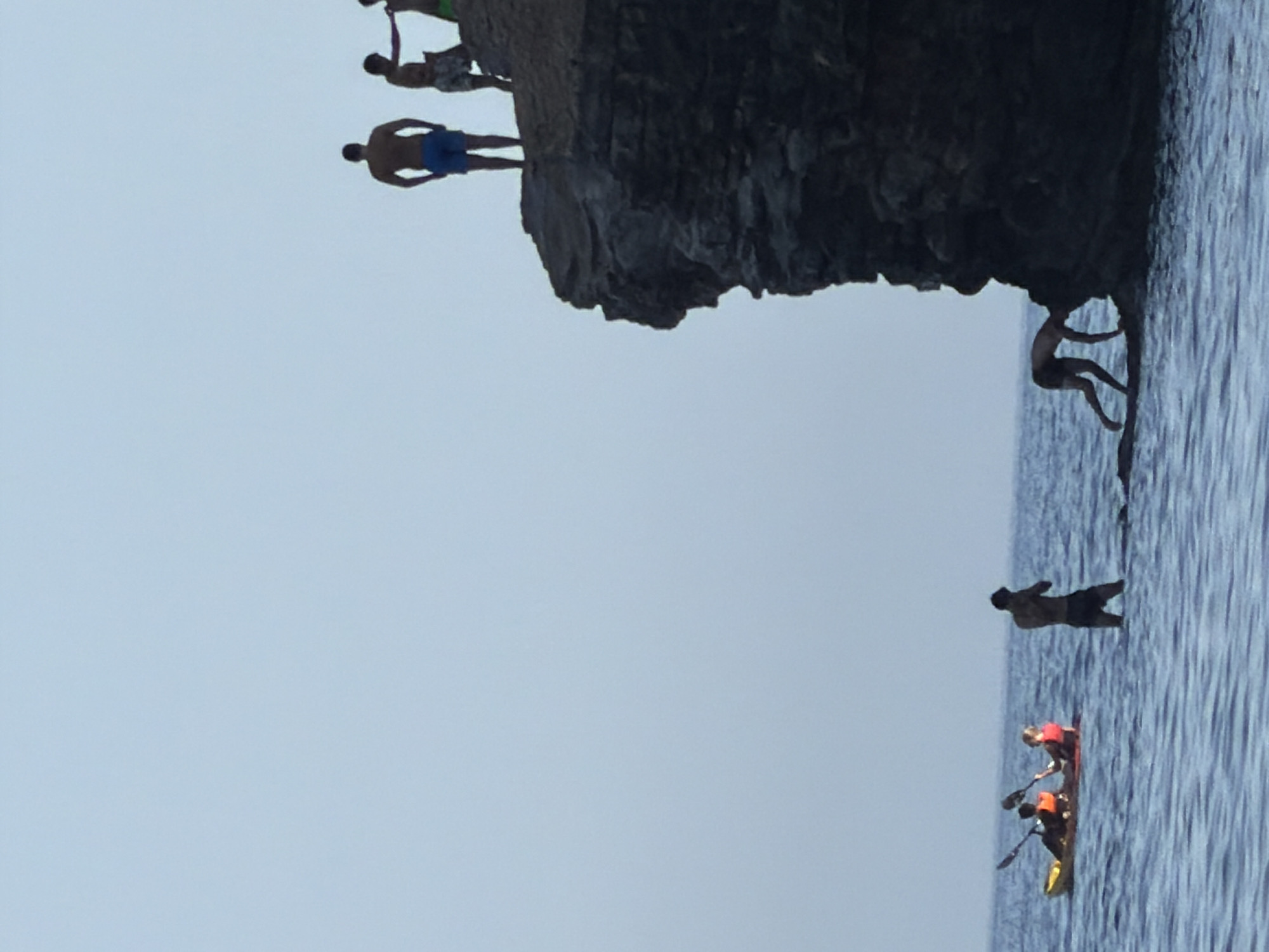 The image shows a cliff face overlooking the Adriatic Sea in Budva, Montenegro. On top of the cliff, several individuals are standing, with one person appearing to be preparing to jump. In the water below, a yellow kayak is being paddled by two people wearing life vests, and another person is standing in the water nearby. The sky is clear and the water is calm.