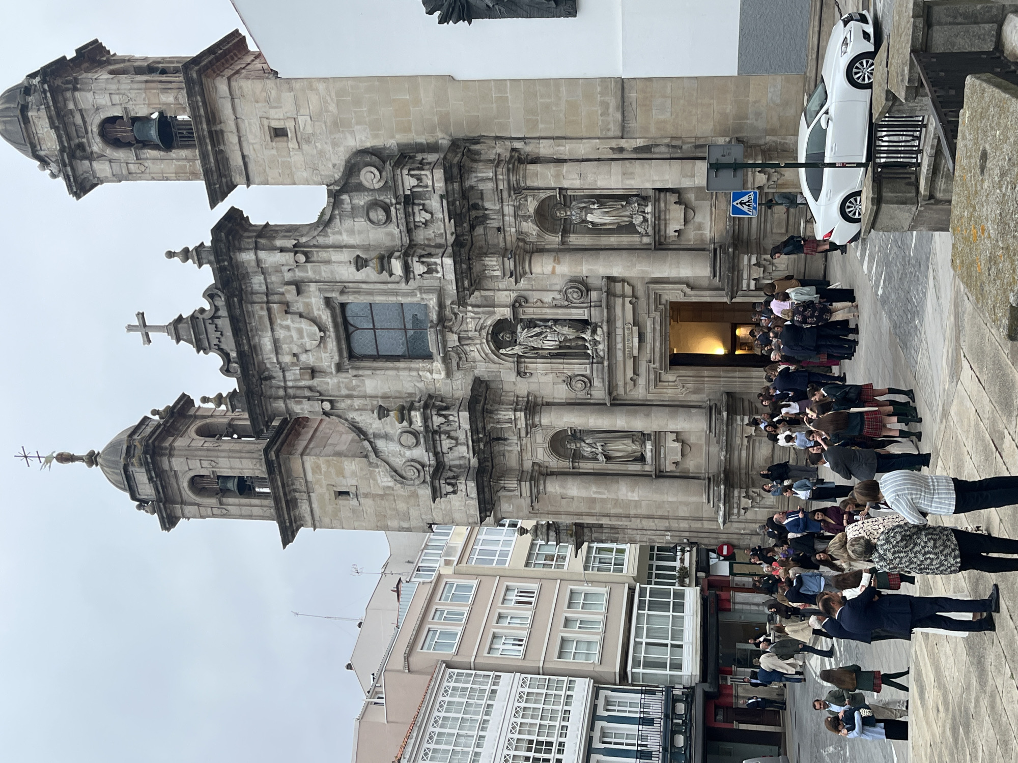 The image shows a large group of people standing in front of a grand, ornate stone building, likely a church or cathedral, with two prominent bell towers. Architectural features include statues in recessed niches on the facade, decorative moldings, and arched windows. The sky is overcast.
Adjacent to the building is a street with parked cars and modern buildings with multiple windows. The people in the foreground appear to be dressed in a mix of formal and casual attire. Their actions include standing, talking, and some appear to be taking photos.
Based on the architecture and general atmosphere, the location appears to be in a city; A Coruña, Spain.