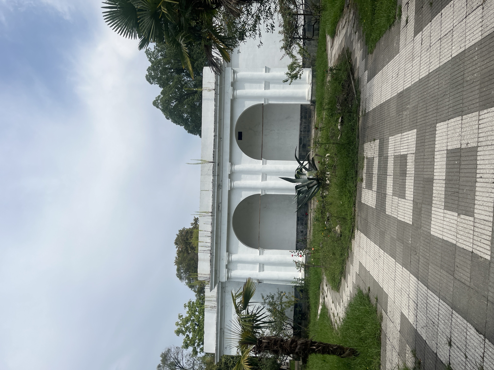 The image depicts an outdoor scene in Sokhumi, Abkhazia, focusing on a white, neoclassical-style building with two arched openings supported by columns. A paved walkway, with a pattern of alternating gray and white rectangular tiles, leads towards the building, curving to the left. Surrounding the walkway and building is a landscape with green grass, various plants, and trees. A palm tree is visible on the left side of the frame, and another is partially visible on the right. The sky is overcast. The building appears to be an architectural structure, possibly a pavilion or part of a larger complex, set within a park or garden environment. There are no people or animals present in the image.