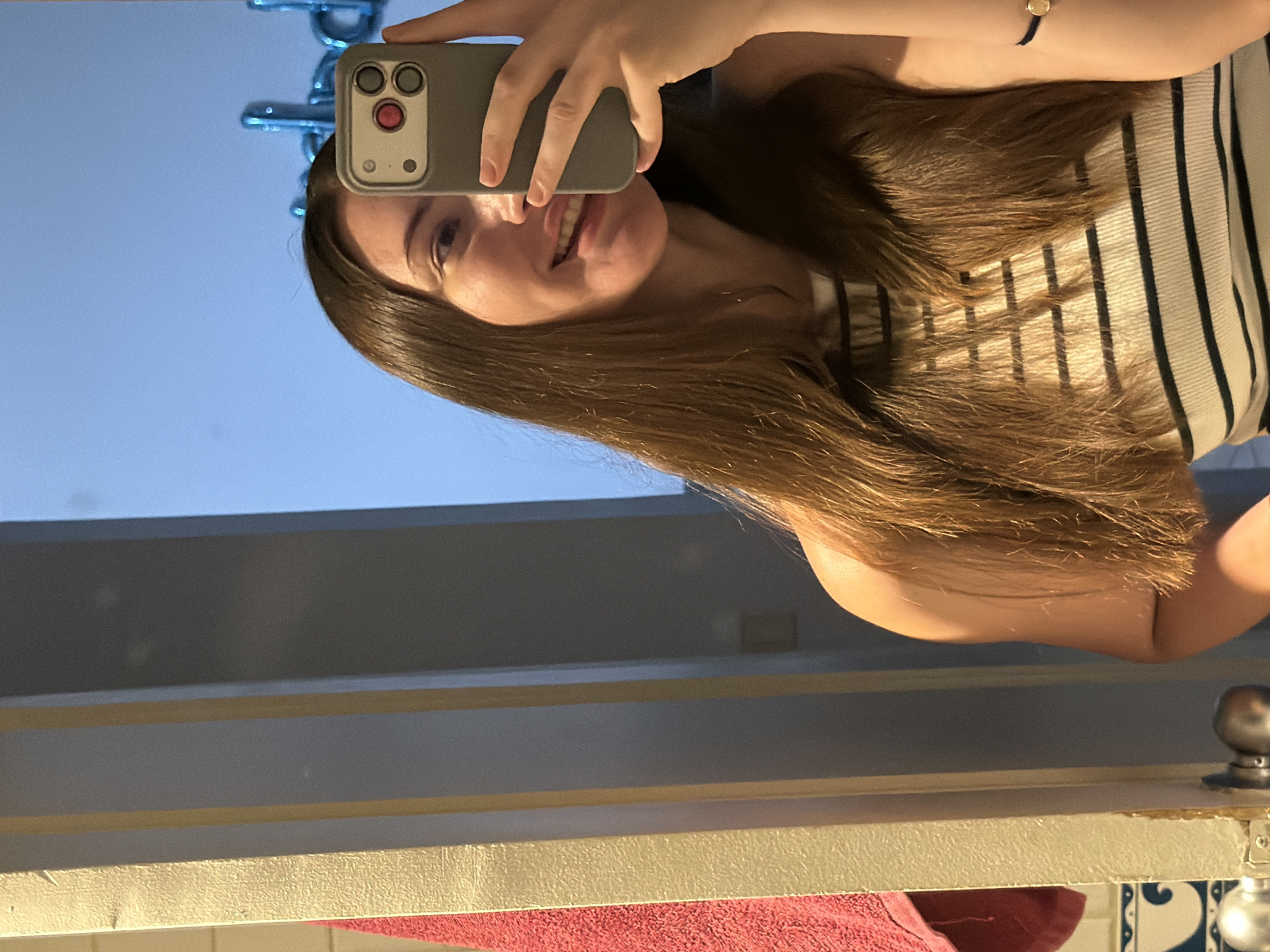 A young Caucasian woman with long, brown hair is taking a selfie in a bathroom. She is wearing a black and white striped tank top. Her left arm is raised, holding a smartphone. A pink towel hangs on a hook to the left of the frame. The background is a light blue wall, possibly a mirror reflecting the sky or a decorated wall. There are blue balloons visible in the reflection. The overall environment appears to be a private residence or hotel bathroom. The image does not provide any specific details to place it in Sharm El Sheikh, Egypt.