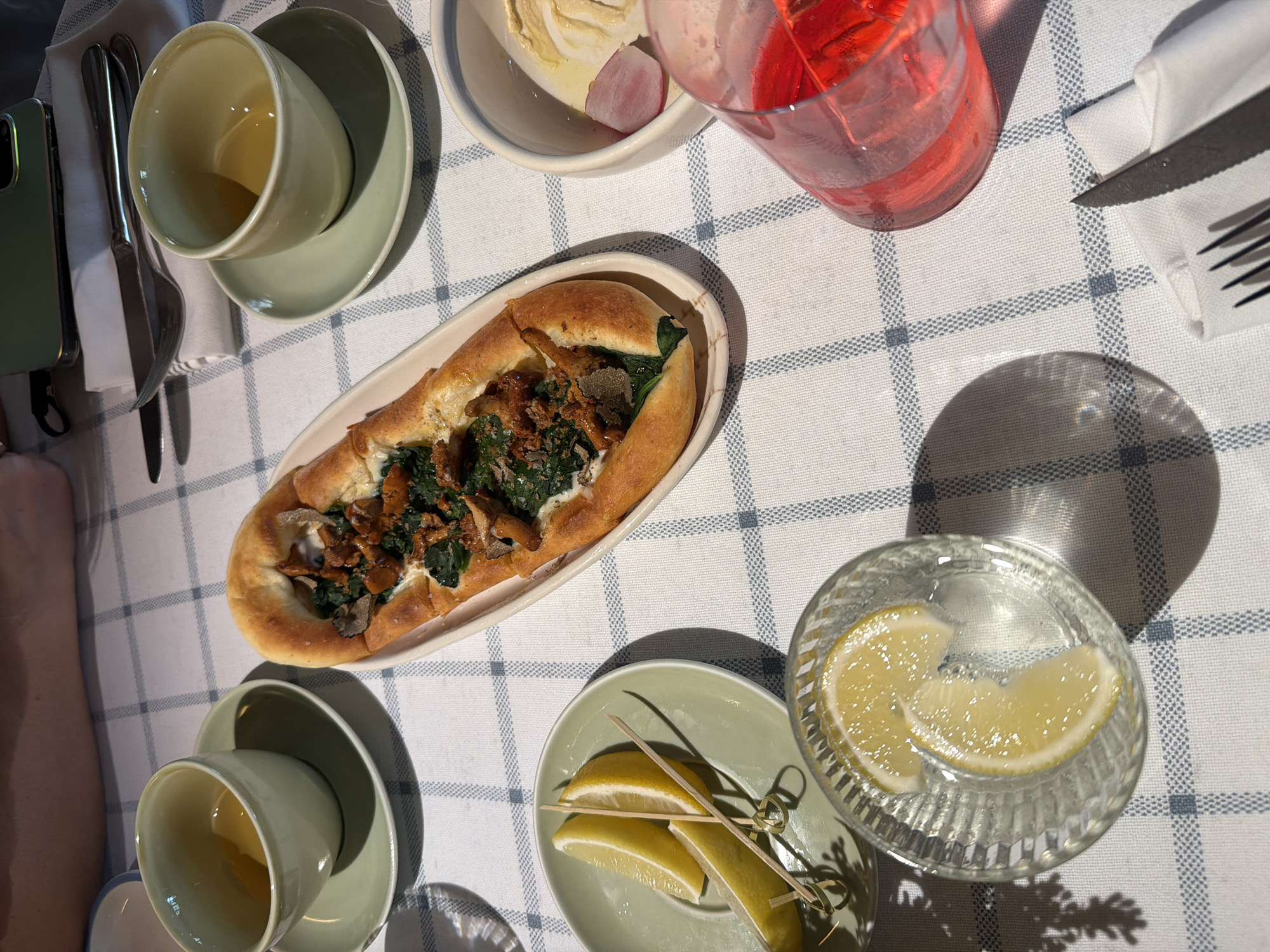 The image shows a table set for a meal, likely in a restaurant in Moscow, Russia, due to the presence of a smartphone with a Russian SIM card visible in the background. The table is covered with a white and blue checkered tablecloth.
On the table, there are several objects:
- A boat-shaped pastry, topped with spinach and what appears to be mushrooms or meat, sits on a light-colored oval plate.
- Two light green cups with saucers, each containing a light yellow liquid, are positioned to the left and right of the pastry.
- A small light green plate holds three lemon wedges skewered with wooden toothpicks.
- A clear glass filled with a red beverage and ice is visible in the lower right corner.
- A small white bowl contains a pale yellow spread and a slice of red radish.
- A knife and fork are placed on a white napkin to the right of the pastry.
- In the background, a person's arm and hand are visible, suggesting someone is seated at the table.
- A smartphone is propped up in the background, its screen illuminated, with what appears to be a charging cable attached.
The lighting suggests an indoor setting, possibly during the day, with shadows cast by the objects. The overall scene conveys a sense of a prepared meal, perhaps a shared appetizer or light dish with drinks.