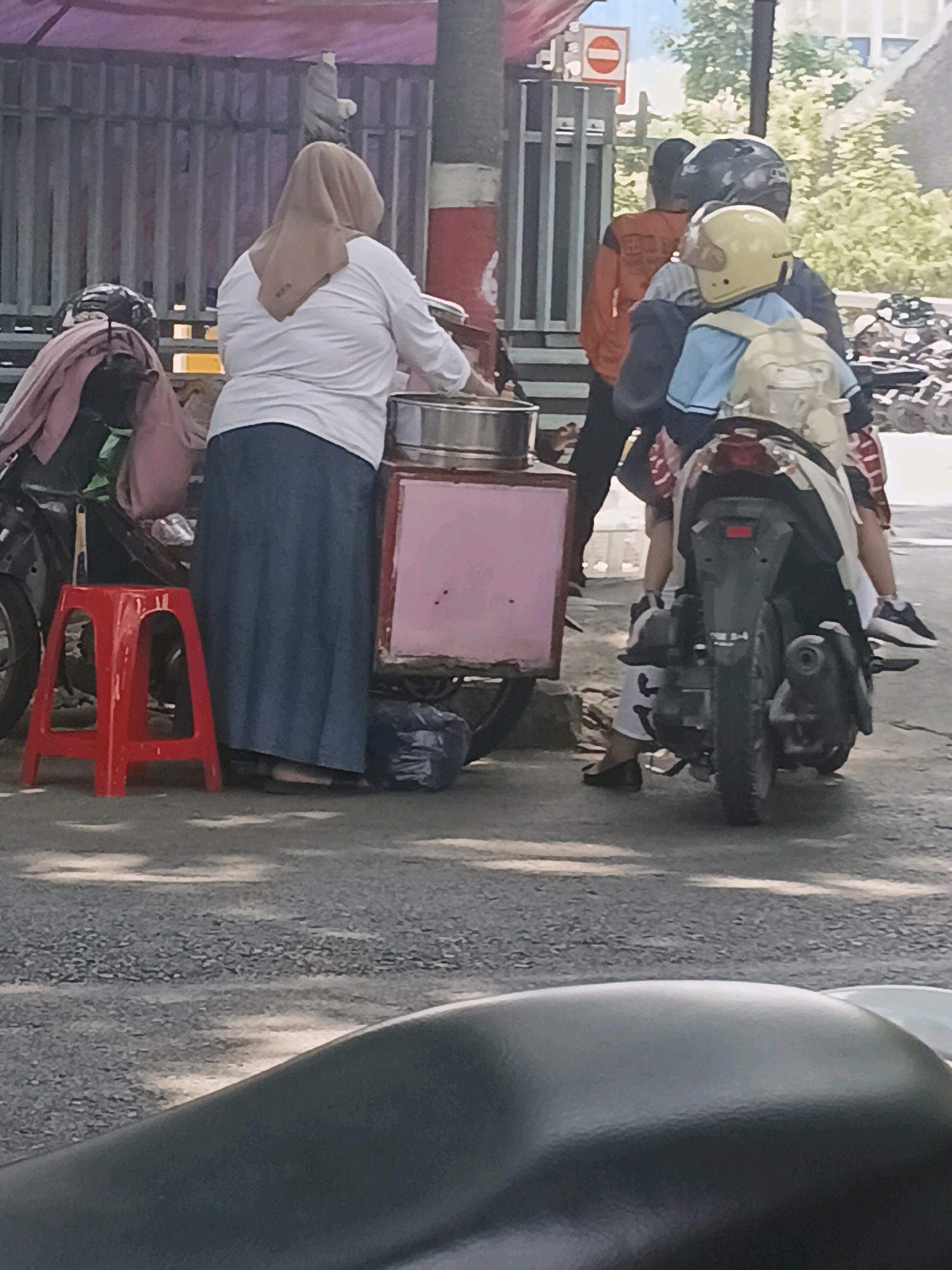 The image shows a street scene in Purwodadi, Indonesia, likely a market or roadside area. A woman wearing a light brown hijab and a white long-sleeved shirt is standing next to a pink mobile cart, possibly selling food. She is wearing a long blue skirt and dark footwear. In front of her is a small red plastic stool. Behind her, a person wearing an orange jacket and a dark helmet is visible, along with another person wearing a light blue shirt and a yellow helmet, both riding a motorcycle. The motorcycle is parked near the cart, and another motorcycle is partially visible on the left side of the frame with clothing draped over it. The background features a metal fence, trees, and a building. The ground is paved with asphalt and gravel.