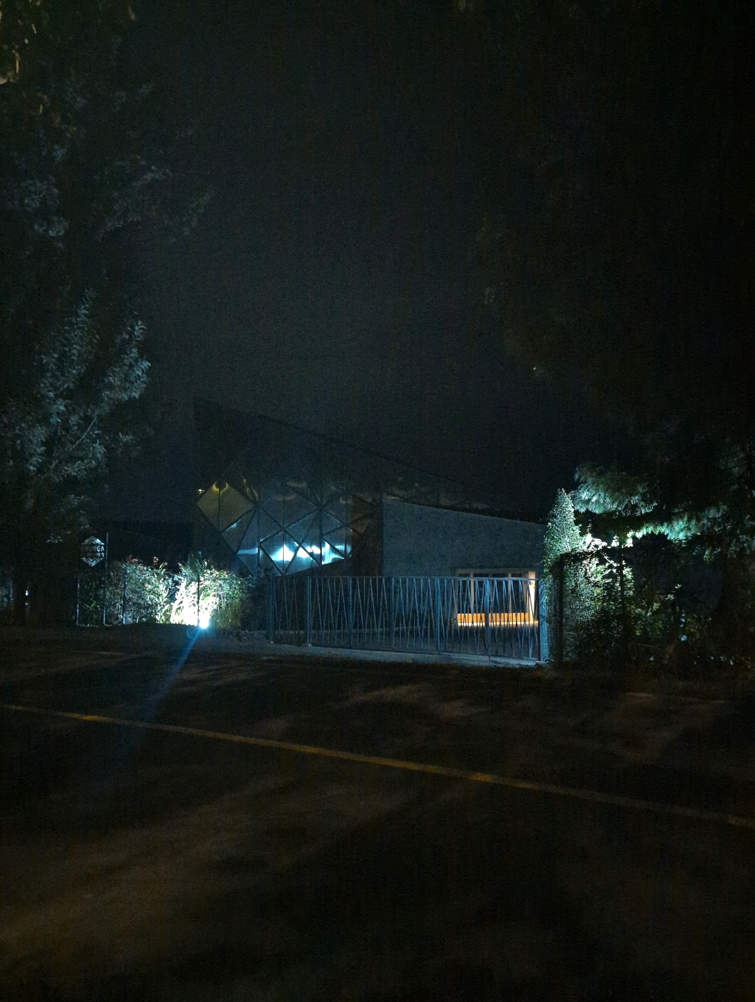 At night in the city of Musanze, Rwanda, a modern, angular building with a glass facade is illuminated from within, casting a blue glow. The structure appears to be made of concrete and glass and is surrounded by trees and dense shrubbery. A metal fence runs along the front of the property, partially obscuring the entrance. A yellow line is visible on the dark asphalt road in the foreground. The scene is dimly lit, with the building and surrounding vegetation being the primary sources of light. The sky is dark and hazy, suggesting the presence of fog or mist.