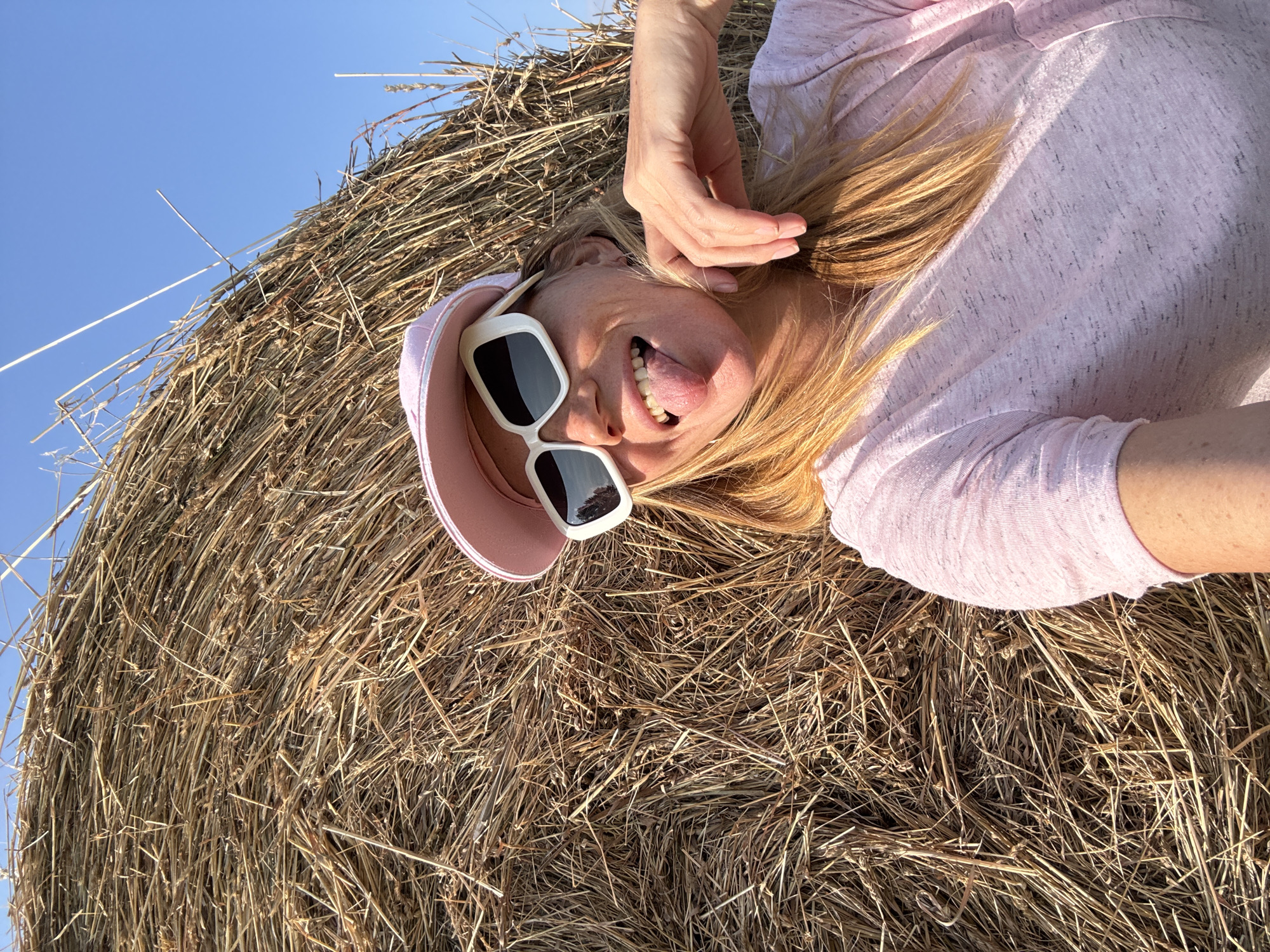 In Yablonovka, Russia, a woman with blonde hair, wearing a pink t-shirt, a pink baseball cap, and white-framed sunglasses, sticks her tongue out at the camera. She is leaning against a large, round bale of hay in a field under a clear blue sky. The hay is dry and golden, with individual stalks clearly visible. The woman's right hand is resting near her chin, and her left arm is visible on the right side of the frame. The sunlight casts shadows across the hay and the woman's face, indicating it is late afternoon.