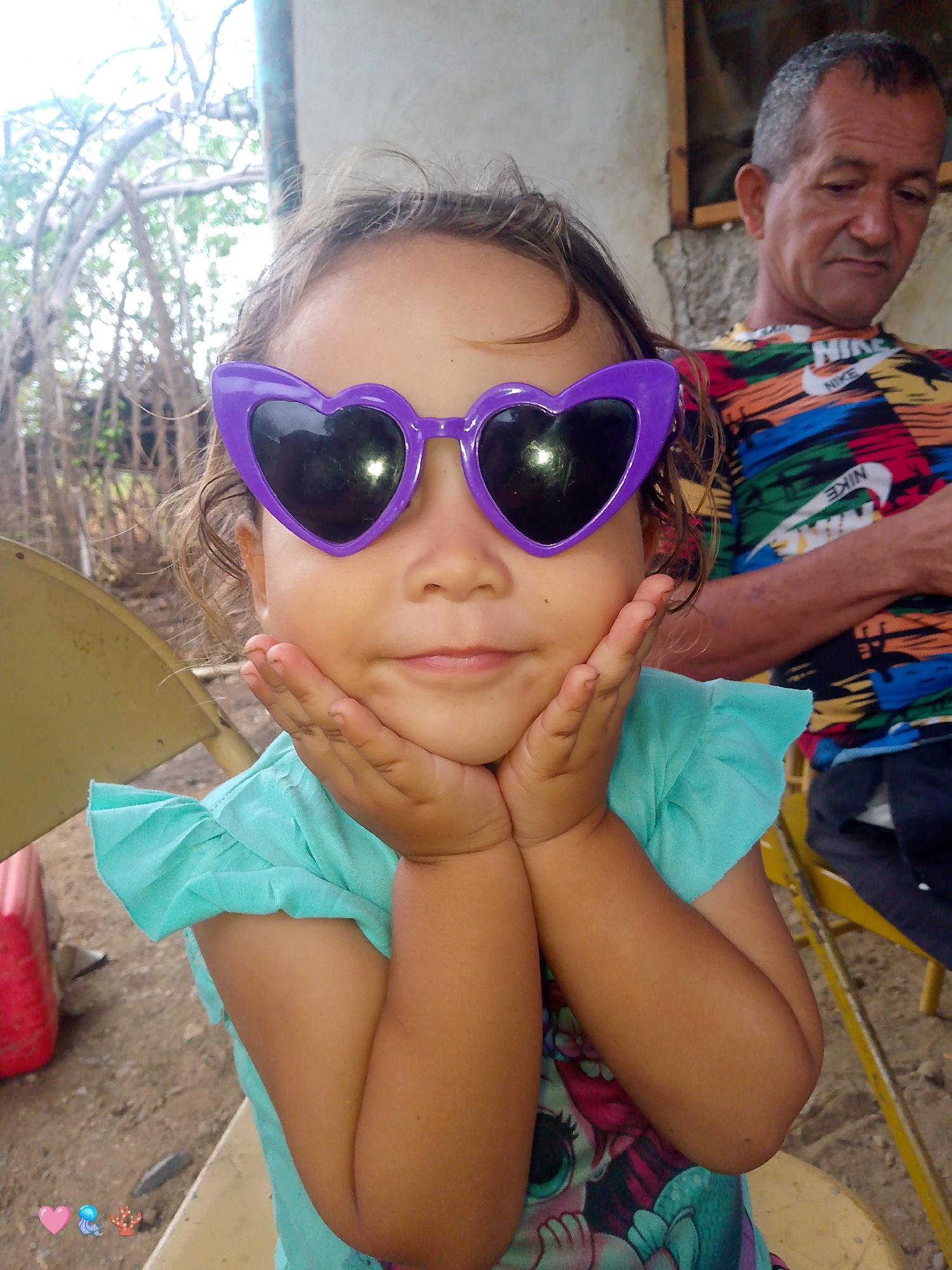 A young girl, wearing oversized purple heart-shaped sunglasses and a turquoise ruffled top, rests her chin on her hands. Behind her, an adult male with short, graying hair and wearing a colorful, patterned t-shirt with a Nike logo is visible. They appear to be outdoors, with a blurred background of trees and structures, suggesting a setting in San Sebastián, Venezuela. The ground is dirt, and a yellow chair is partially visible.