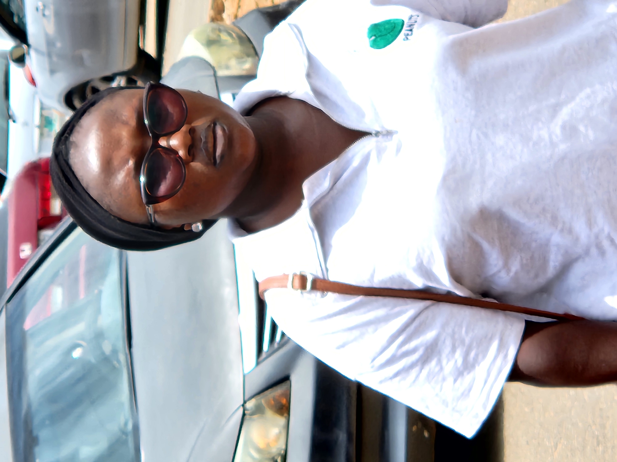 A close-up, waist-up shot shows a Black woman wearing a white polo shirt with a green logo and the word "PEANUTS" underneath, and a black headscarf. She is also wearing sunglasses with brown lenses and silver earrings. She has a brown strap from a bag across her chest. In the background, two cars are visible; one is dark gray, and the other is light silver. The scene appears to be outdoors, possibly in a parking lot or street in Jos, Nigeria, given the lighting and background elements. The woman is looking directly at the camera, and her expression is neutral. The image quality is somewhat soft, with visible noise and minor blurring.
