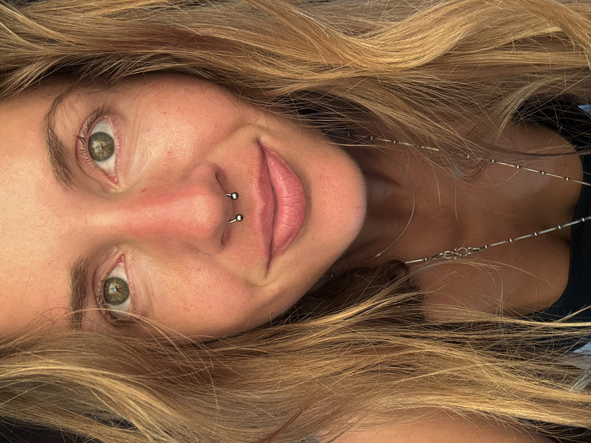 This is a close-up, medium shot, eye-level portrait of a young Caucasian woman with long, wavy blonde hair. She is facing the camera, and her head is slightly tilted to her left. Her skin is fair with freckles and appears sun-kissed with a reddish hue around her nose and cheeks. She has green eyes, arched eyebrows, and visible eyelashes. She is wearing a septum piercing, which is a small horseshoe-shaped metal ring. Her lips are full and appear to be a light pink color. She is wearing a simple, thin silver necklace with small beads. The background is dark and out of focus, suggesting an indoor or dimly lit environment. The lighting is warm and casts a glow on her face.
