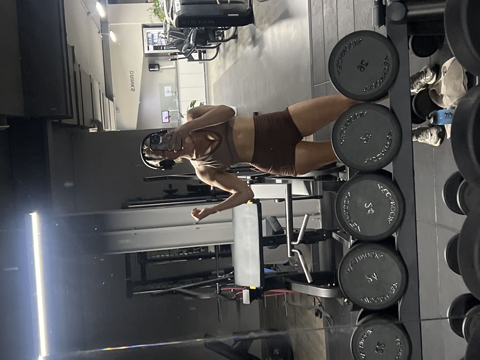 The image shows a person in a gym, taking a selfie in a mirror. The person is wearing a sports bra and shorts, and appears to be in good shape. They are wearing headphones, and their left arm is extended towards the camera. In the background, there are weight racks with dumbbells, gym equipment, and a sign that reads "DRINKS" in reverse. The gym appears to be well-lit and modern. There are also reflections of the gym in the mirror, including the person's legs and more gym equipment. The image is taken from a low angle, looking up at the person. The text "TECHNOGYM" is visible on some of the dumbbells, along with the weight "45". The overall mood of the image is one of fitness and self-care. The time of day is not specified, but the lighting suggests it could be daytime or evening. There is no indication of weather. The location context is Altötting, Germany.