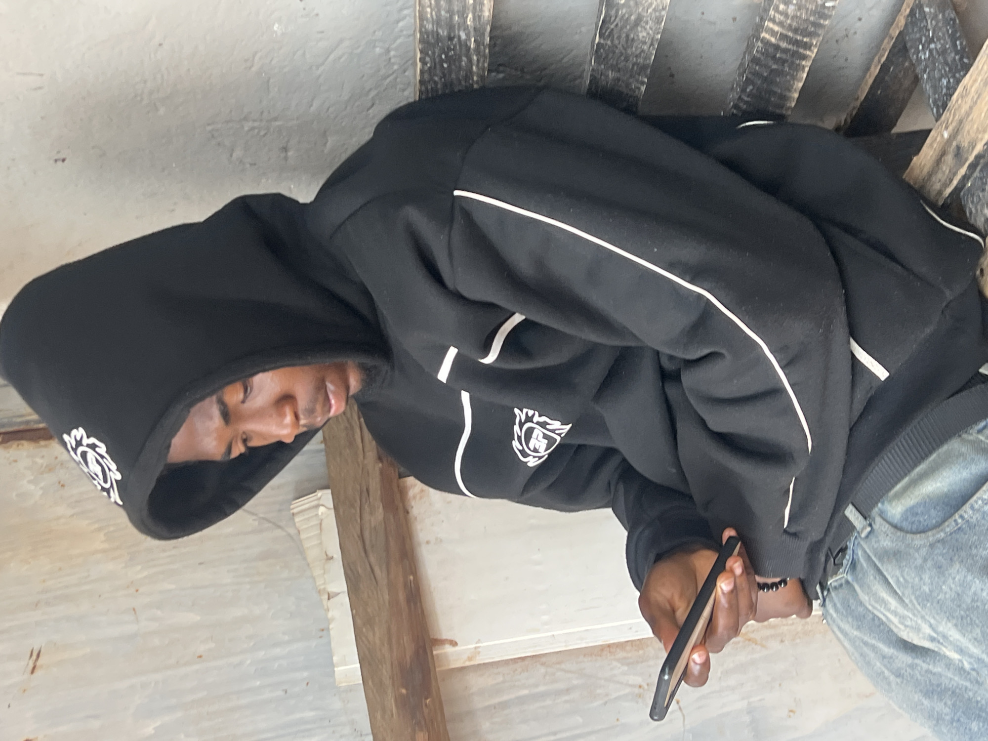 A young Black male is sitting on a wooden bench, wearing a black hoodie with white trim and a logo on the left breast, and light blue denim jeans. He is looking down at his mobile phone, which he is holding in his left hand. The background consists of a corrugated metal wall and some wooden planks. The scene appears to be outdoors in a somewhat rustic setting. It is not possible to determine the specific location within the city of Durimi, Nigeria from the image alone.