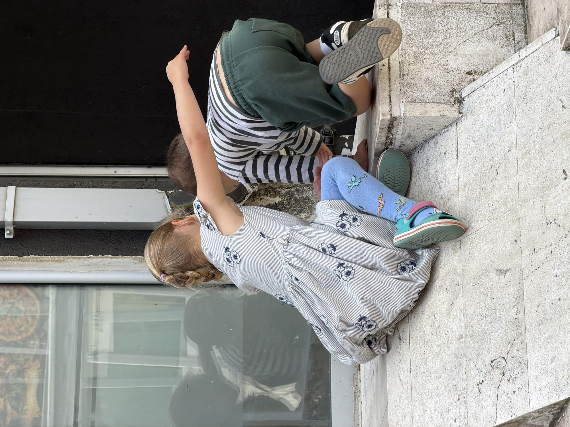 Two children are on a stone staircase in an urban setting. One child wears a striped shirt, green shorts, and black and white shoes and is bending over a ledge. The other child, a girl, wears a striped dress with floral print, blue patterned leggings, and teal sandals. A glass window with a white frame is behind the girl, reflecting an image of the scene. The background includes a dark-colored wall. The staircase is made of light-colored stone and has various cracks and textures. The location is possibly in Budva, Montenegro.