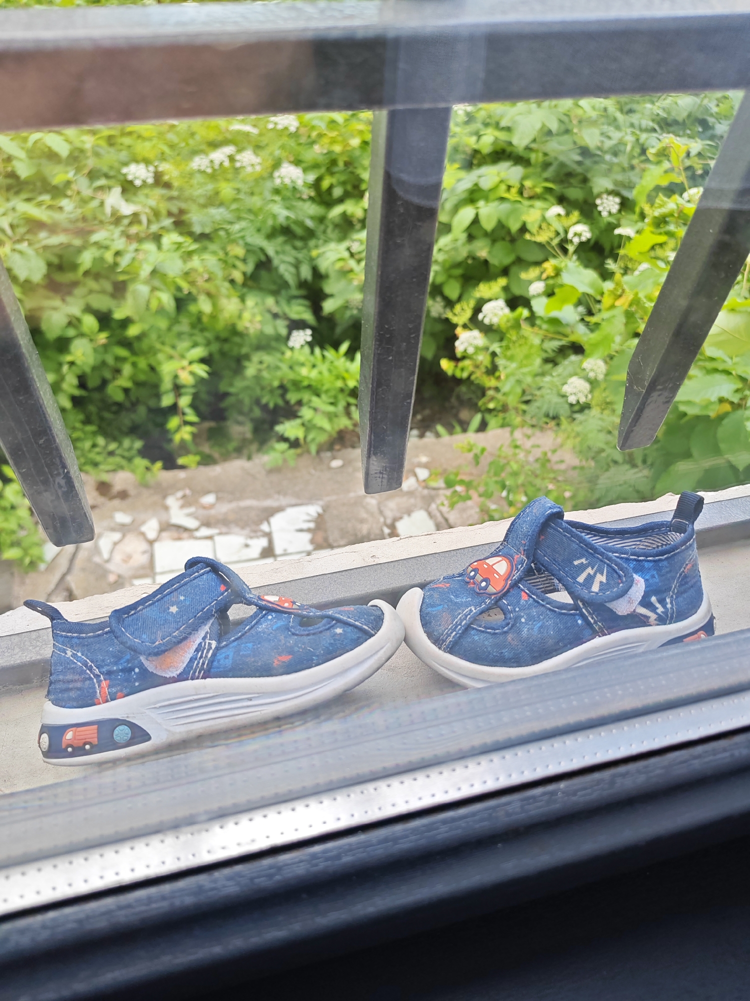 The image shows a pair of children's blue canvas shoes with a white sole, featuring a pattern of red trucks and white stars. The shoes are placed on a windowsill, with the left shoe closer to the camera and the right shoe slightly behind and to the right. The background consists of lush green foliage with white flowers, suggesting an outdoor setting with vegetation. The scene is framed by what appears to be a dark metal railing or bars, possibly part of a balcony or window guard. The lighting suggests daytime. The location is identified as Vanadzor, Armenia.