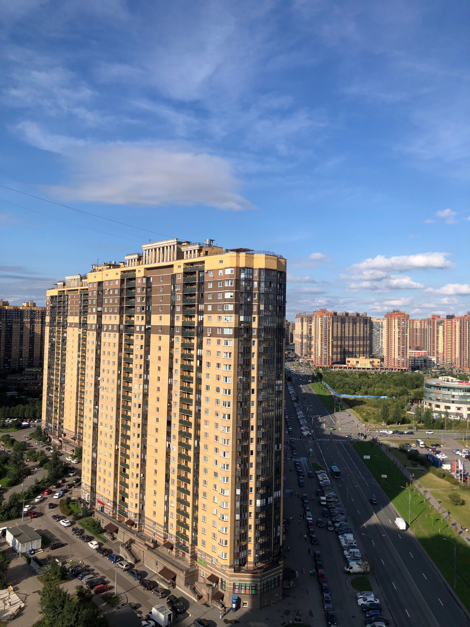 This is an aerial photograph of a residential area in Lavriki, Russia, characterized by tall, multi-story apartment buildings. The prominent building in the foreground is a large, cream-colored structure with numerous windows and dark brown vertical accents. Its facade is punctuated by balconies, many of which are enclosed with glass. The building has a decorative cornice at the top.
Beyond this building, the landscape reveals a dense collection of similar high-rise apartments stretching into the distance. These buildings are primarily colored in shades of orange and cream, with dark vertical elements and multiple windows. They are arranged along a wide, multi-lane road that runs through the scene.
The road is lined with parked cars on both sides, indicating a densely populated area. Traffic appears to be moving along the road, with several vehicles visible, including a bus. Green spaces, including trees and grassy areas, are interspersed between the buildings and along the roadside, suggesting a planned urban development.
The sky above is a bright blue with scattered white clouds, indicating fair weather. Power lines are faintly visible against the sky in the upper portion of the image. The perspective suggests the photograph was taken from a very high vantage point, likely from another tall building or a drone.