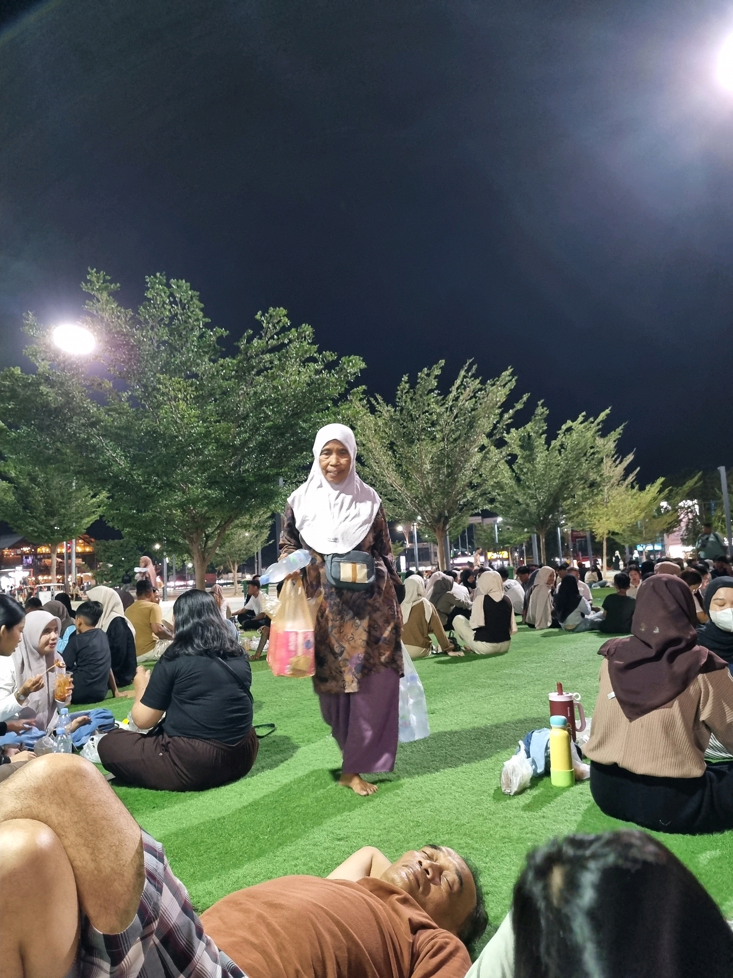 The image depicts an outdoor gathering at night in Tanamodindi, Indonesia. A diverse group of people are seated on a vibrant green artificial turf. In the foreground, a man wearing a brown t-shirt is lying down, his head resting on a pillow. To his right, a person wearing a brown hijab and a beige sweater is also seated. A woman in a white hijab and a patterned brown and purple dress walks in the midground, carrying plastic bags and a water bottle. She is smiling as she looks towards the camera. Behind her, several groups of people are seated, some in conversation, others appearing to eat or relax. Lush green trees with dense foliage are visible in the background, illuminated by overhead lighting. The scene suggests a community event or a relaxed social gathering in a park or public space within the city.