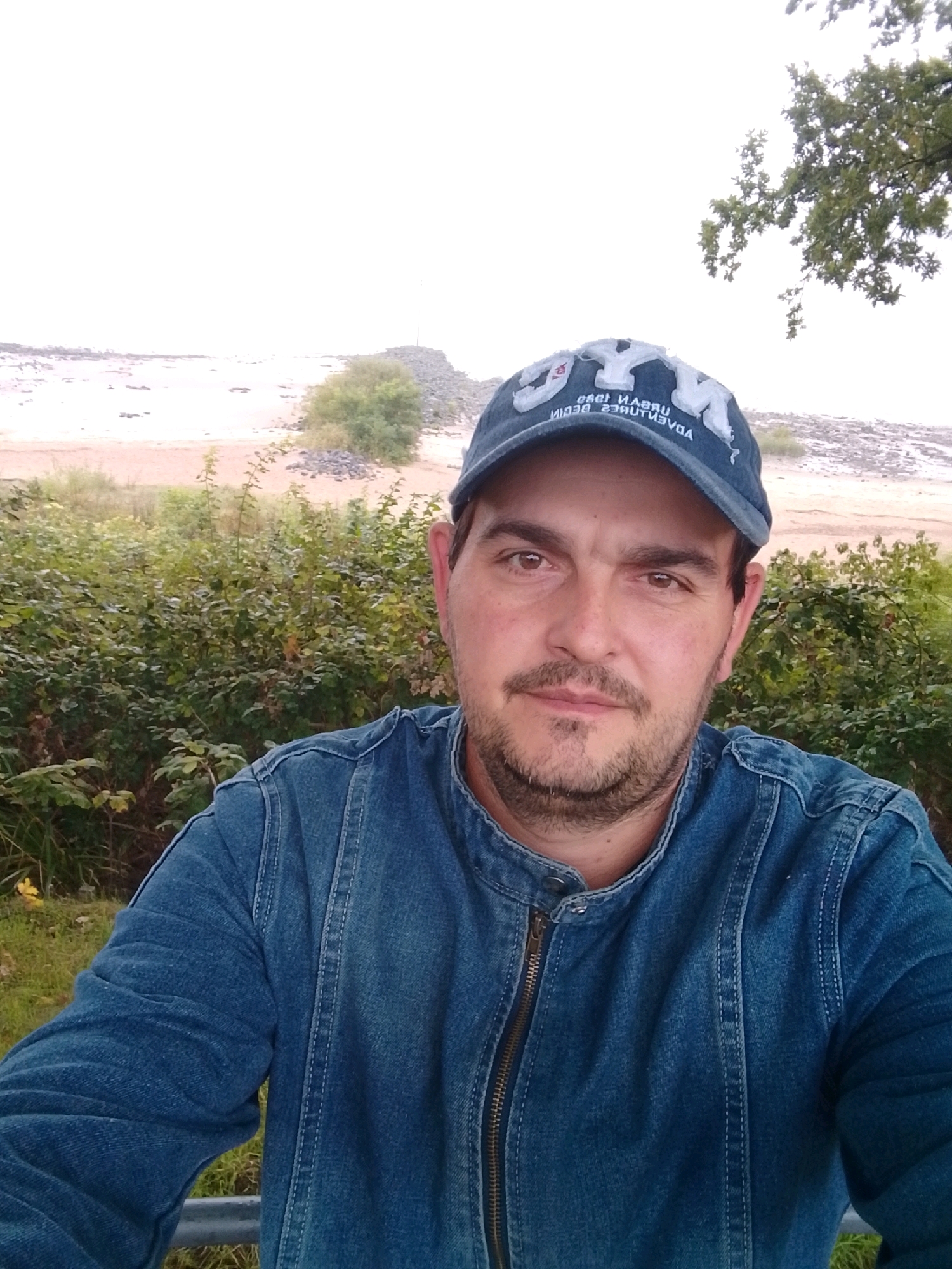 A man wearing a blue denim jacket and a blue baseball cap is captured in a close-up, outdoor, selfie-style photograph. The man has brown eyes, short brown hair, and a light beard and mustache. His cap is emblazoned with white lettering, some of which is reversed due to the nature of a selfie. Behind him, a landscape unfolds, featuring dense green foliage, a sandy or gravelly embankment, and a hazy sky. The overall impression is one of a casual outdoor setting, possibly a park or natural area. The image does not contain sufficient information to determine the specific location within the city of Wedel, Germany.