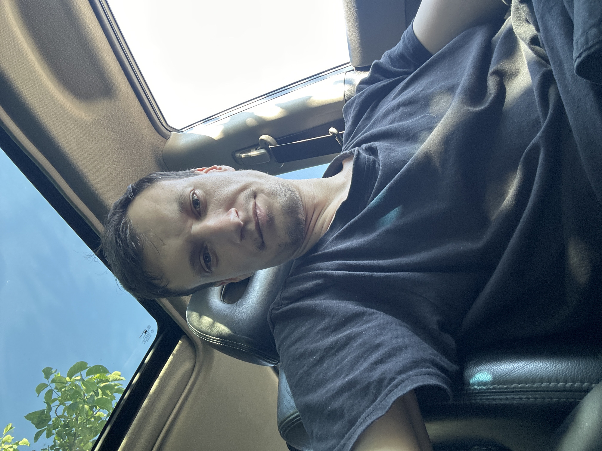 In Shatava, Ukraine, a male individual is seated in the driver's seat of a vehicle, looking towards the camera. The individual is wearing a dark t-shirt. Sunlight streams through the vehicle's sunroof, illuminating the interior and casting shadows on the man's face and clothing. The sunroof is open, revealing a clear blue sky with some green foliage visible. The interior of the car features black leather seats and a beige headliner. The overall scene suggests an outdoor, daytime setting, possibly during a journey.