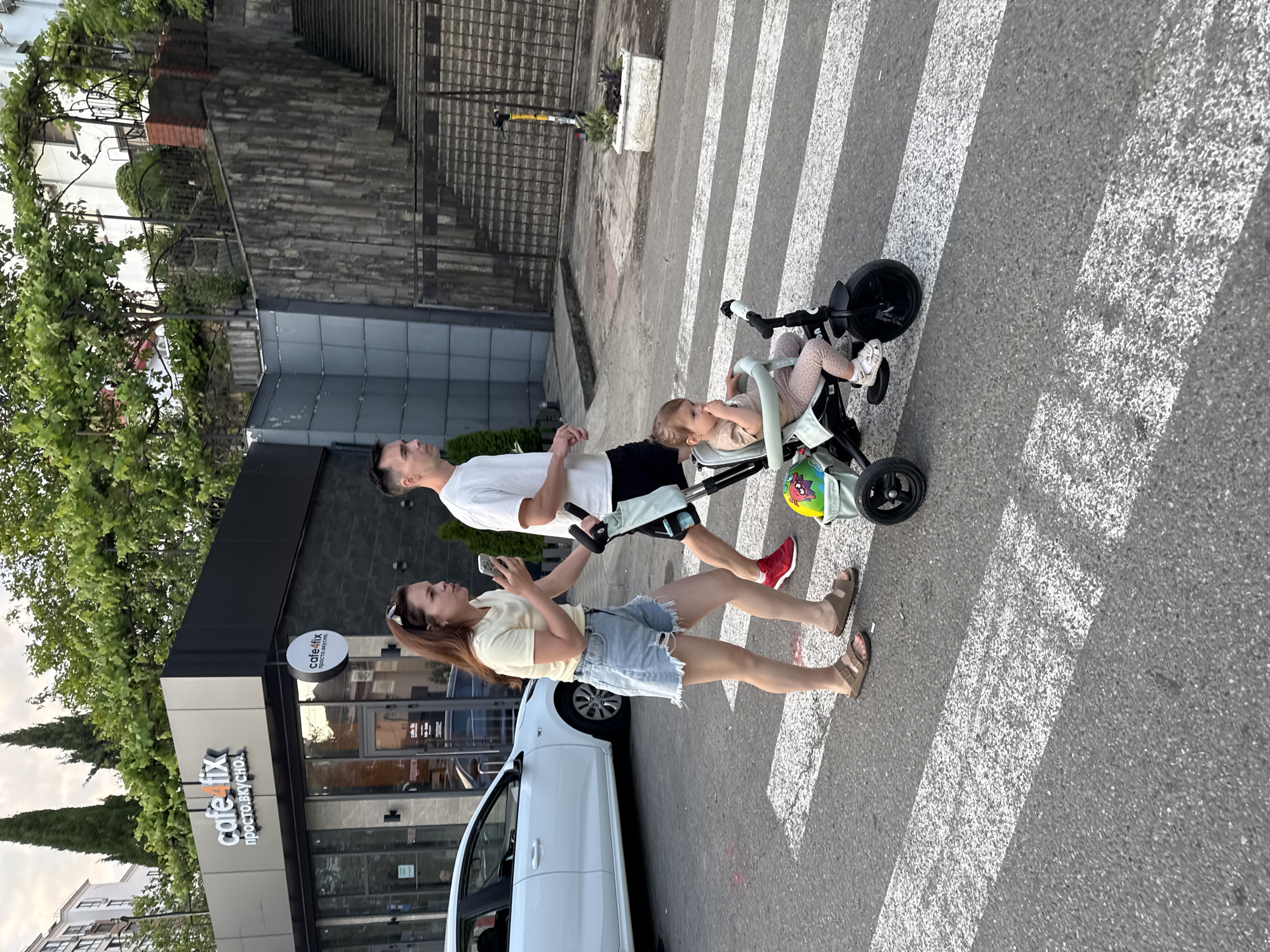 The image depicts a family crossing a pedestrian street in Sochi, Russia. A woman, a man, and a young child are visible. The woman, wearing a yellow t-shirt and denim shorts, walks on the left, holding her phone. The man, in a white t-shirt and black shorts, walks beside her, pushing a tricycle with the child in it. The child is seated in the tricycle, wearing a light-colored outfit and socks. The tricycle is green and black, with a colorful bag attached to the back. The background features a building with a sign that reads "cafe fix." To the right, a stone wall and a staircase are visible, with greenery and trees surrounding the area. The setting appears to be an urban environment with a pedestrian crossing marked by white stripes on the asphalt.