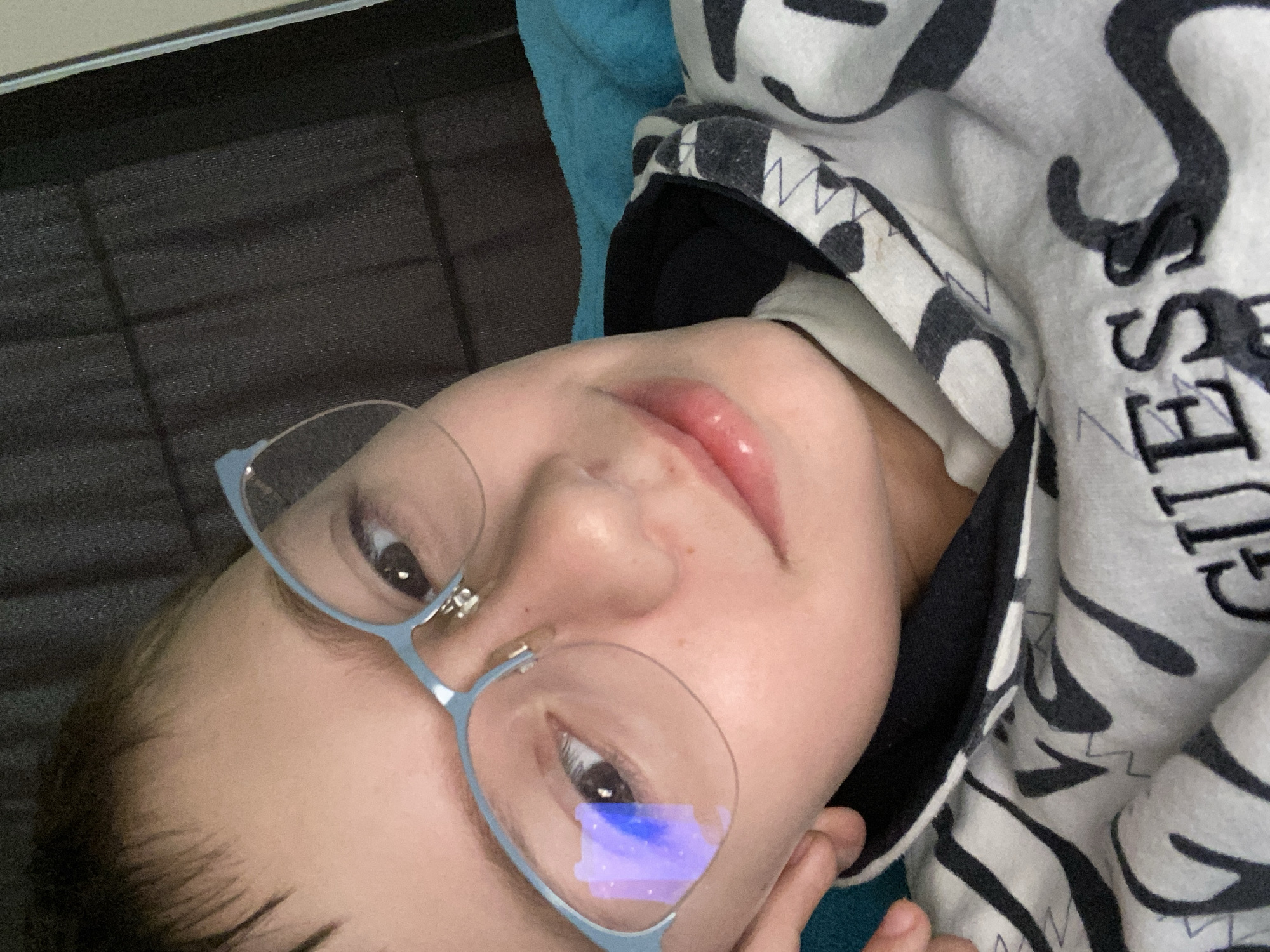 A young person with fair skin is lying down, facing the camera at an angle. They are wearing light blue, round eyeglasses. The person's lips have a glossy appearance. They are wearing a hooded sweatshirt with a white and black zebra stripe pattern and the word "GUESS" printed on it. A dark gray patterned surface is in the background, along with a blue blanket or pillow. There is no indication that this image was taken in Burgas, Bulgaria.