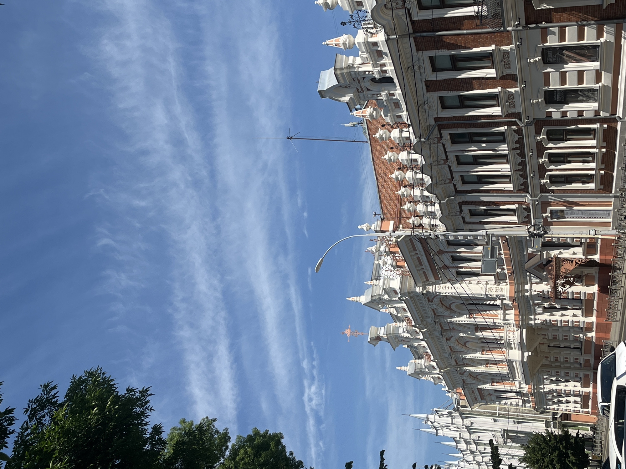 The image captures a street view in Krasnodar, Russia, focusing on the ornate architecture of buildings lining the street. The buildings feature a distinct style with intricate decorative elements, contrasting brickwork, and white stucco moldings. The sky above is a vibrant blue with wispy cirrus clouds. A street lamp is visible in the mid-ground, and tree branches with green foliage are present on the left side of the frame, partially obscuring the sky. Several cars are parked along the street in the foreground. The overall scene depicts a sunny day in an urban environment with a focus on historical architecture.