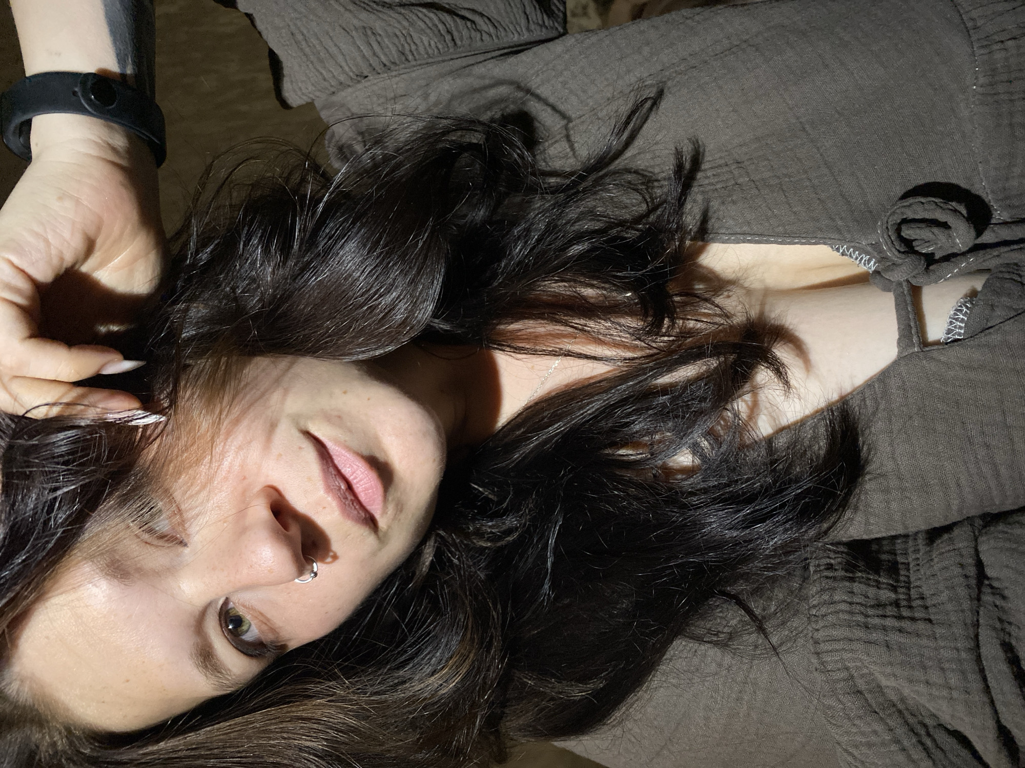 The image shows a close-up of a woman's face and upper body. She has long, dark, wavy hair, with some strands falling over her face. She is wearing a dark, textured top with a tie closure at the neckline, revealing a glimpse of a bra underneath. She has a nose ring on her left nostril and is wearing a black watch on her left wrist. Her skin appears fair with freckles. The lighting suggests an indoor setting, possibly a home environment, with soft, directional light casting shadows. There is no indication of the specific location within Chaikovsky, Russia, in the image itself.