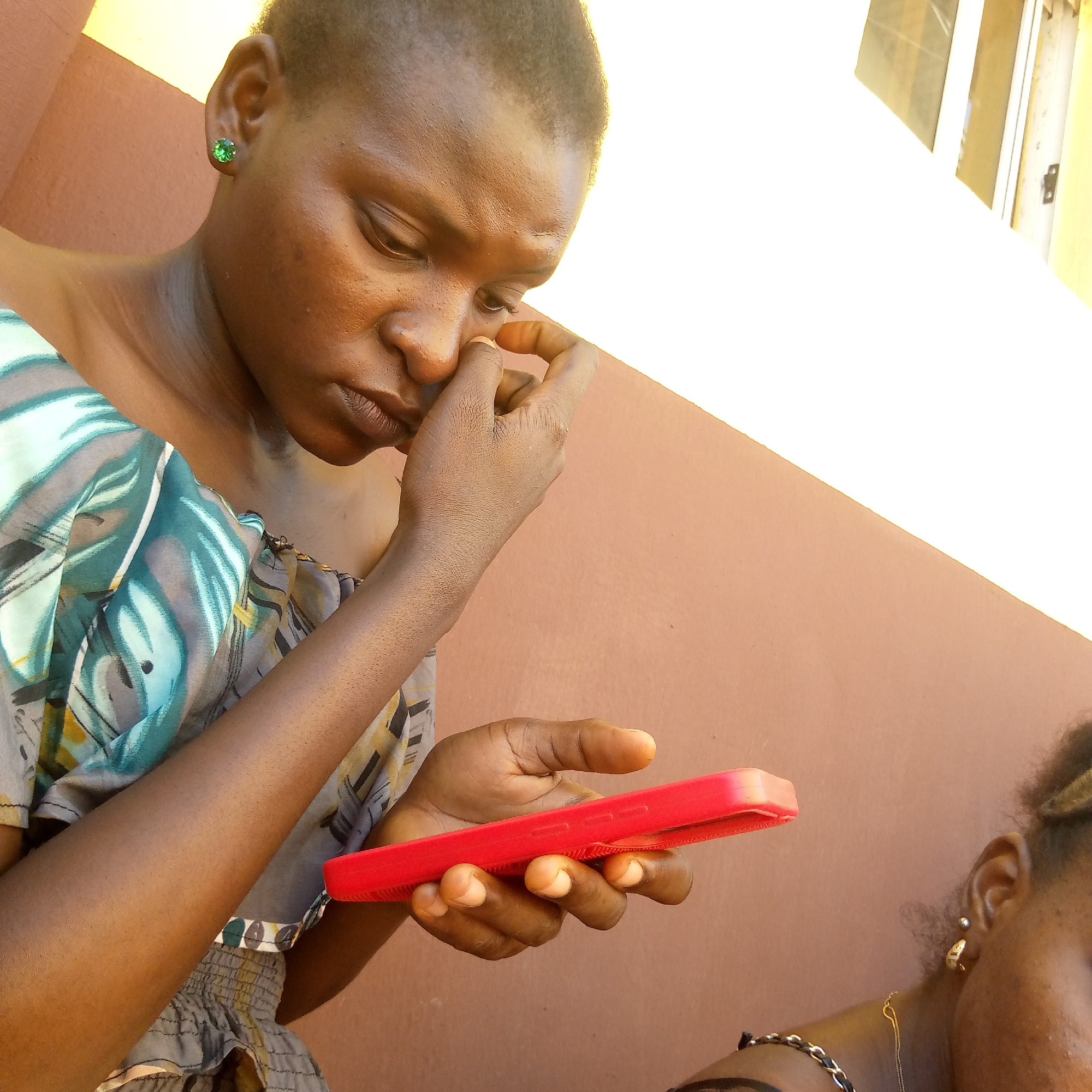 A medium shot captures a young, dark-skinned woman holding a red smartphone while appearing preoccupied with her nose. She is wearing a light, patterned top, and a green stud earring is visible in her left ear. Another woman is partially visible in the lower right corner. They are situated next to a wall. Considering the prompt, the location is in Ukwu-Abwa, Nigeria.