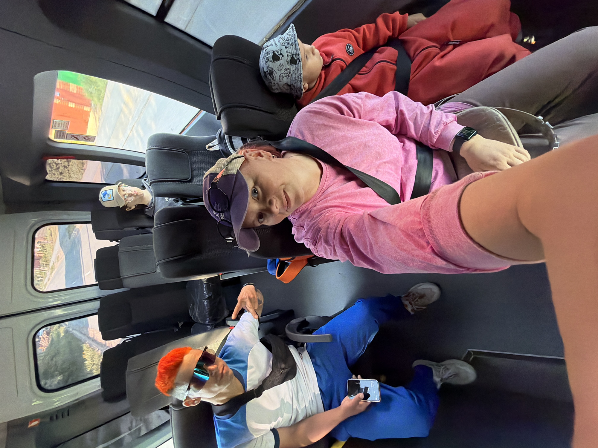 Here's a description of the image:
Inside a vehicle, possibly a van or bus, are several people. In the foreground, a person with orange hair and sunglasses is sitting and holding a mobile phone. They are wearing a blue and white top and blue trousers. Behind them is a woman wearing a pink top, a purple hat, and a black wristwatch. Next to her is a child in a red hoodie and a hat patterned with skulls. All three appear to be wearing seatbelts. The rows of seats in the vehicle are mostly empty, and outside the windows, there is a view of buildings and trees. The vehicle appears to be in Chaikovsky, Russia.