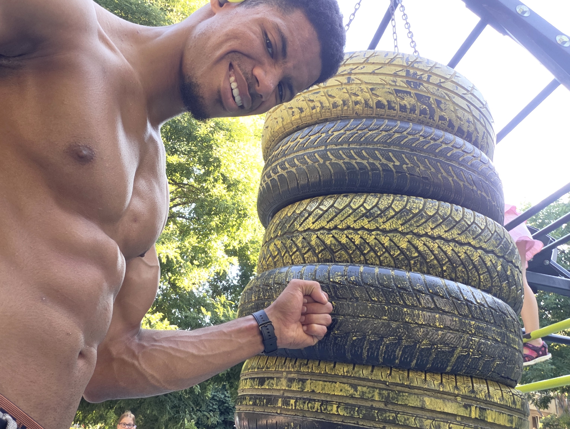 A shirtless man with defined muscles smiles into the camera, leaning his head against a stack of tires. The tires, painted yellow and black, hang from chains. The man is wearing a black watch on his left wrist. In the background, green trees and a bright sky are visible. A child in a pink dress and sandals stands on a metal structure behind the tires. The scene appears to be outdoors, possibly in a park or playground setting. No specific Varna landmarks are identifiable.