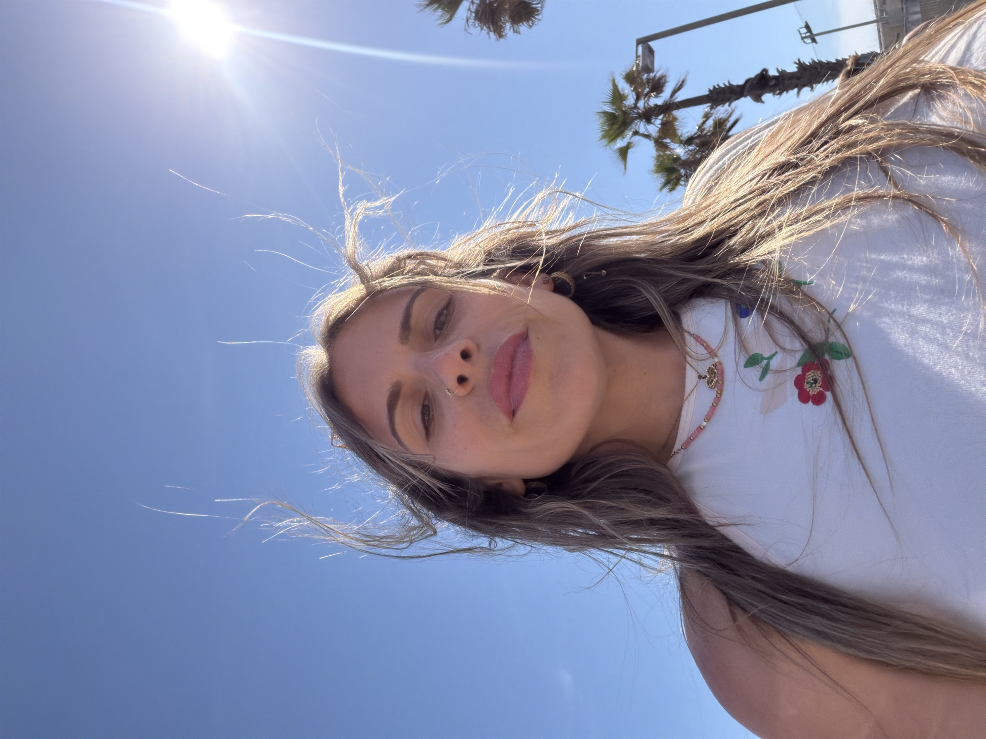 A tilted portrait shot captures a young woman from a low angle, looking up towards the bright blue sky on a sunny day in Barcelona, Spain. The sun is intensely bright in the upper left, casting lens flare and strongly backlighting her long, light brown hair, which appears windblown with individual strands highlighted against the sky.
The woman's face is centered in the frame, and she gazes directly at the viewer with a neutral to slightly confident expression. Her lips are lightly colored, and she wears a nose ring and earrings. She is dressed in a white top, partially visible, featuring embroidered floral designs in red and green. A delicate necklace is also seen around her neck.
In the upper background, several palm trees and a utility pole or street light are silhouetted against the clear sky, confirming an outdoor, warm-weather setting. The strong sunlight and clear skies indicate it is likely midday or early afternoon. There is no visible text in the image.