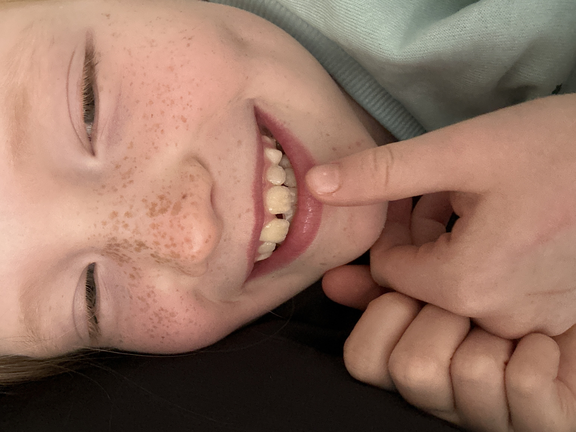 A close-up photograph depicts a young person lying down. Their eyes are mostly closed. Their face is freckled, and they are smiling, revealing their teeth. A hand is positioned near their mouth, with a finger touching their lower lip. The person is wearing a light-colored sweatshirt. The background is dark, possibly clothing or bedding. The location of Уральское, Russia, cannot be verified from the image.