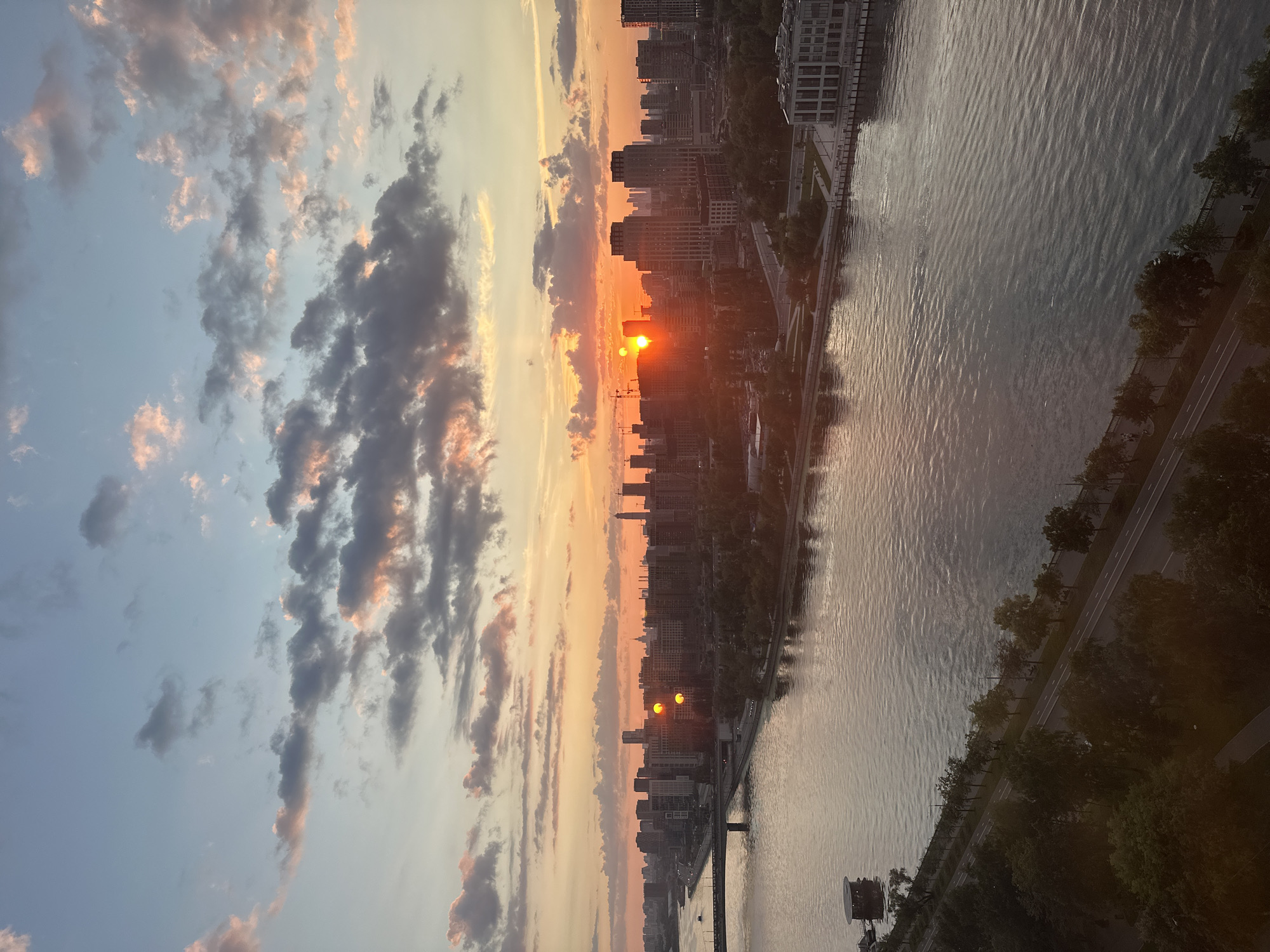 The image depicts a wide river flowing through a modern cityscape at sunset. The sky is filled with dramatic clouds, tinged with orange and pink hues from the setting sun, which is positioned low on the horizon and partially obscured by buildings. A dense urban skyline dominates the middle ground, characterized by numerous tall, contemporary buildings. A bridge spans the river in the foreground, connecting two sides of the city. Along the riverbanks, there are tree-lined promenades and walkways. The water reflects the colors of the sky and the surrounding architecture. The scene captures the transitional light of dusk over an urban environment.