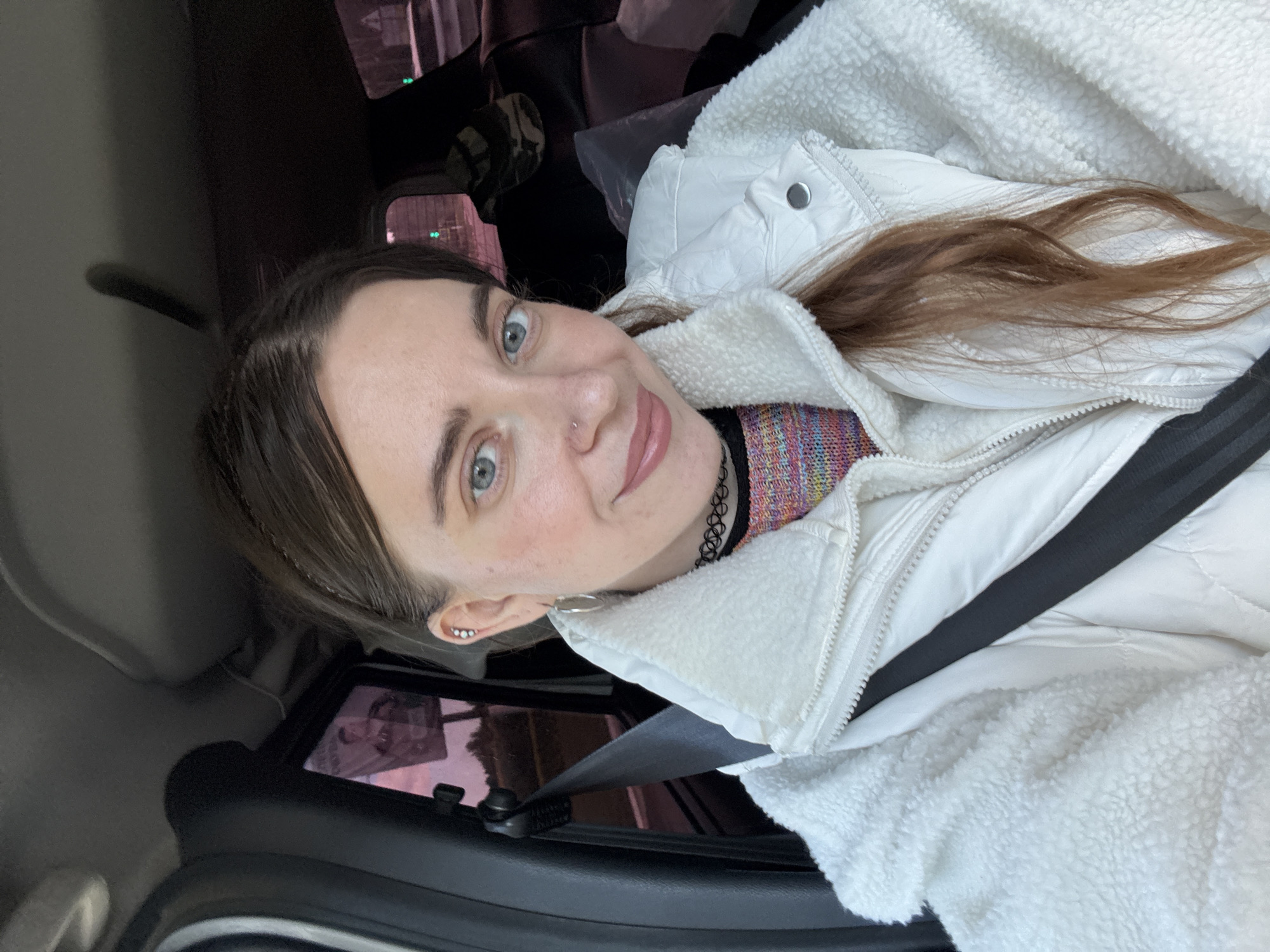The image shows a young woman with blue eyes, brown hair, and a nose ring, taking a selfie in a car. She is wearing a white fuzzy jacket over a colorful patterned shirt and a black choker necklace. The seatbelt is visible across her chest. The background is blurred, but suggests a nighttime setting with some lights visible through the car windows. The woman is smiling and appears relaxed. The photo is taken from a slightly tilted angle, making it appear as though the car is moving. There is no visible text in the image.