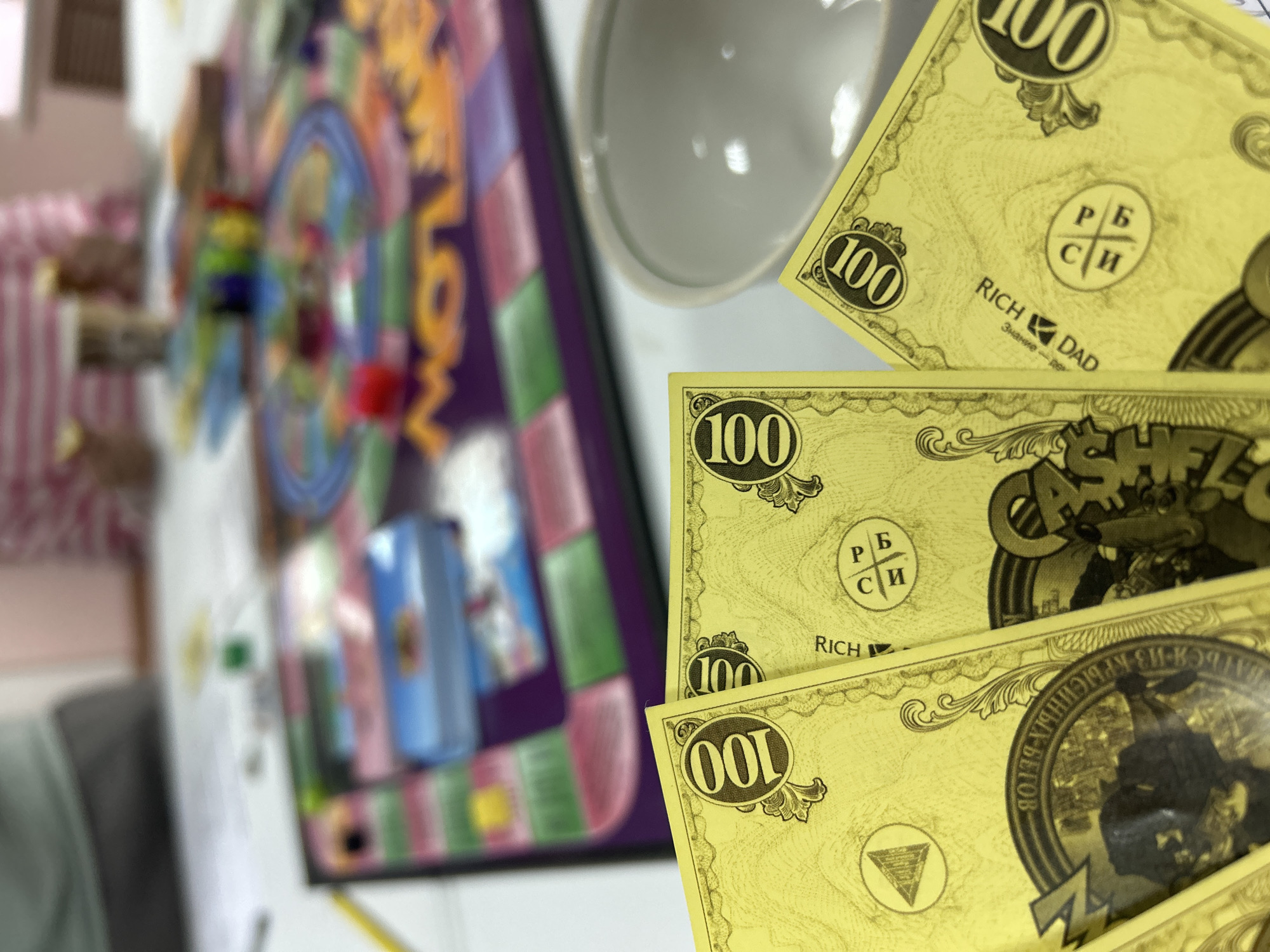 The image captures a close-up, angled view of a board game setup, likely in Chaikovsky, Russia. In the sharp foreground, several yellow play money bills, each marked "100", are fanned out on a white surface. The bills feature the text "RICH DAD" and include Cyrillic characters such as "Р Б", "С И", "Знание – де", and "КРЫСИНЫХ-Ы БЕГОВ", strongly suggesting currency from the "Cashflow" board game, which is based on Robert Kiyosaki's "Rich Dad Poor Dad" principles and the concept of escaping the "rat race" (крысиных бегов).
Behind the money, a white bowl is partially visible. The background is blurred, showing a colorful board game board, with elements resembling game pieces and squares. The word "LOW" is discernible on the board, further indicating the "Cashflow" game. The overall scene suggests an indoor recreational activity, likely a board game session. The lighting appears to be ambient indoor light.
