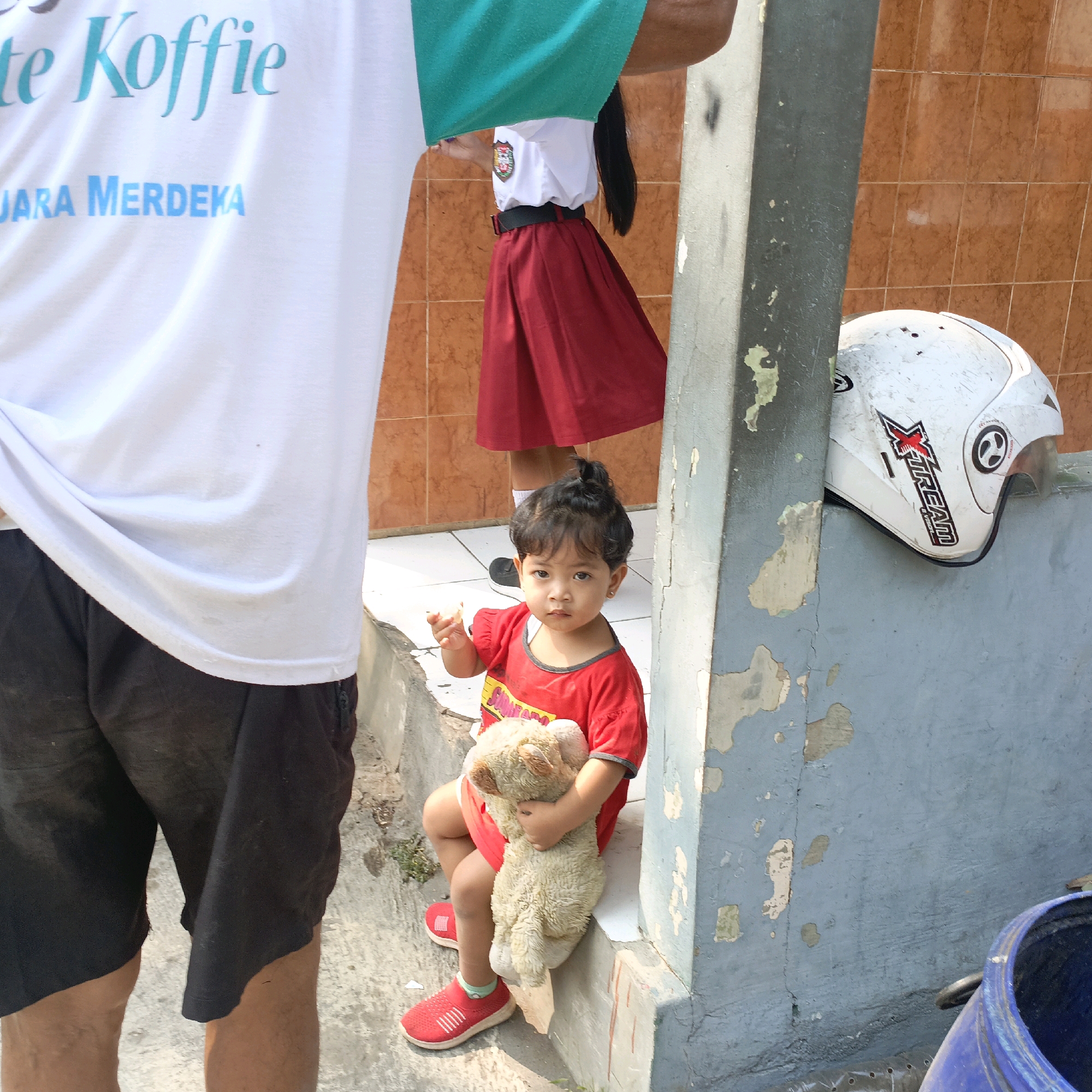 The image shows a scene outside a building, likely in Salatiga, Indonesia, given the context provided. In the foreground, a young child, possibly a girl, is seated on a ledge, clutching a stuffed animal. The child wears a red t-shirt and red shoes with white stripes. To the left of the child, a person, mostly out of frame, is wearing a white t-shirt with text printed on it, including "te Koffie" and "JUARA MERDEKA". This person also wears black shorts. Behind the child, another person in a school uniform, consisting of a white shirt and a red skirt, stands partially visible. The background features a tiled wall and a light-colored concrete structure. A white motorcycle helmet with red and black markings that read "XTREAM" is propped against the concrete structure. The environment appears to be a public or residential area, with a blue plastic container visible in the lower right corner. The lighting suggests it is daytime.